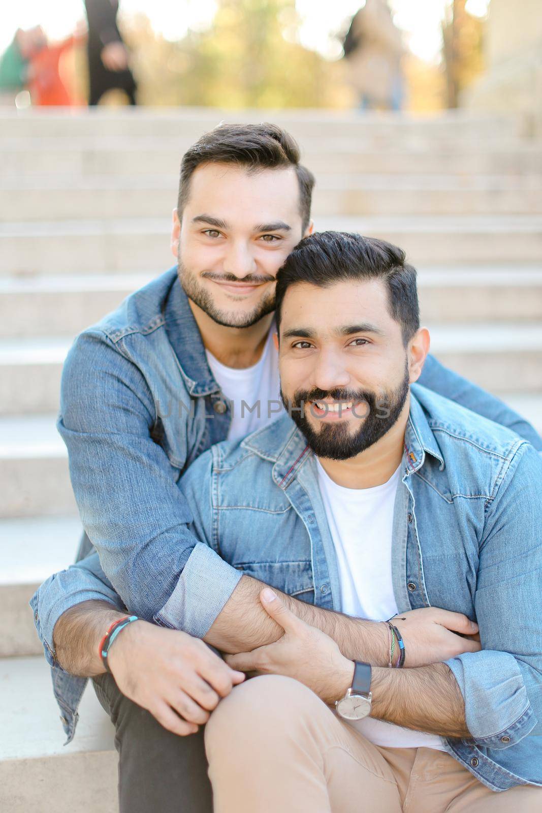Caucasian handsome european young gays sitting on concrete stairs and hugging, wearing jeans shirts. Concept of same sex couple and lgbt.