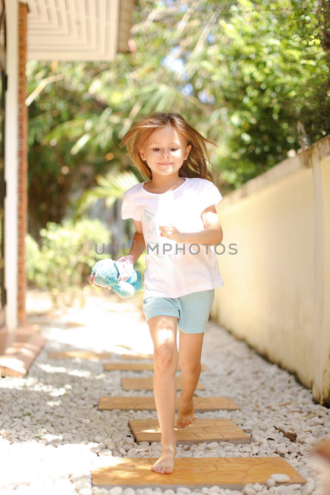 Little caucasian female kid running barefoot on white gravel near exotic hotel. Concept of vacation with kid in Thailand, Phuket.