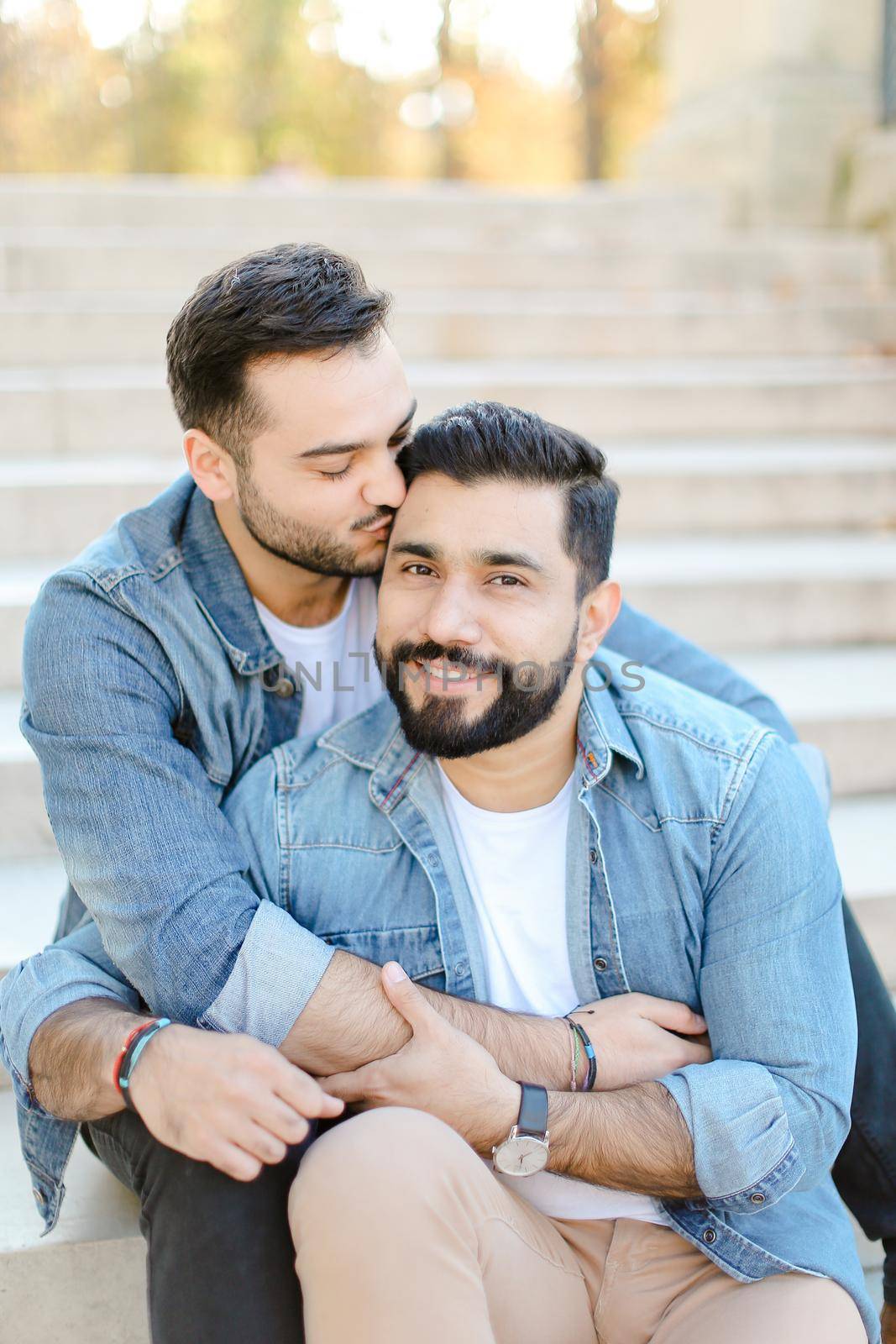 Caucasian young gays sitting on concrete stairs and kissing, wearing jeans shirts. Concept of same sex couple and lgbt.
