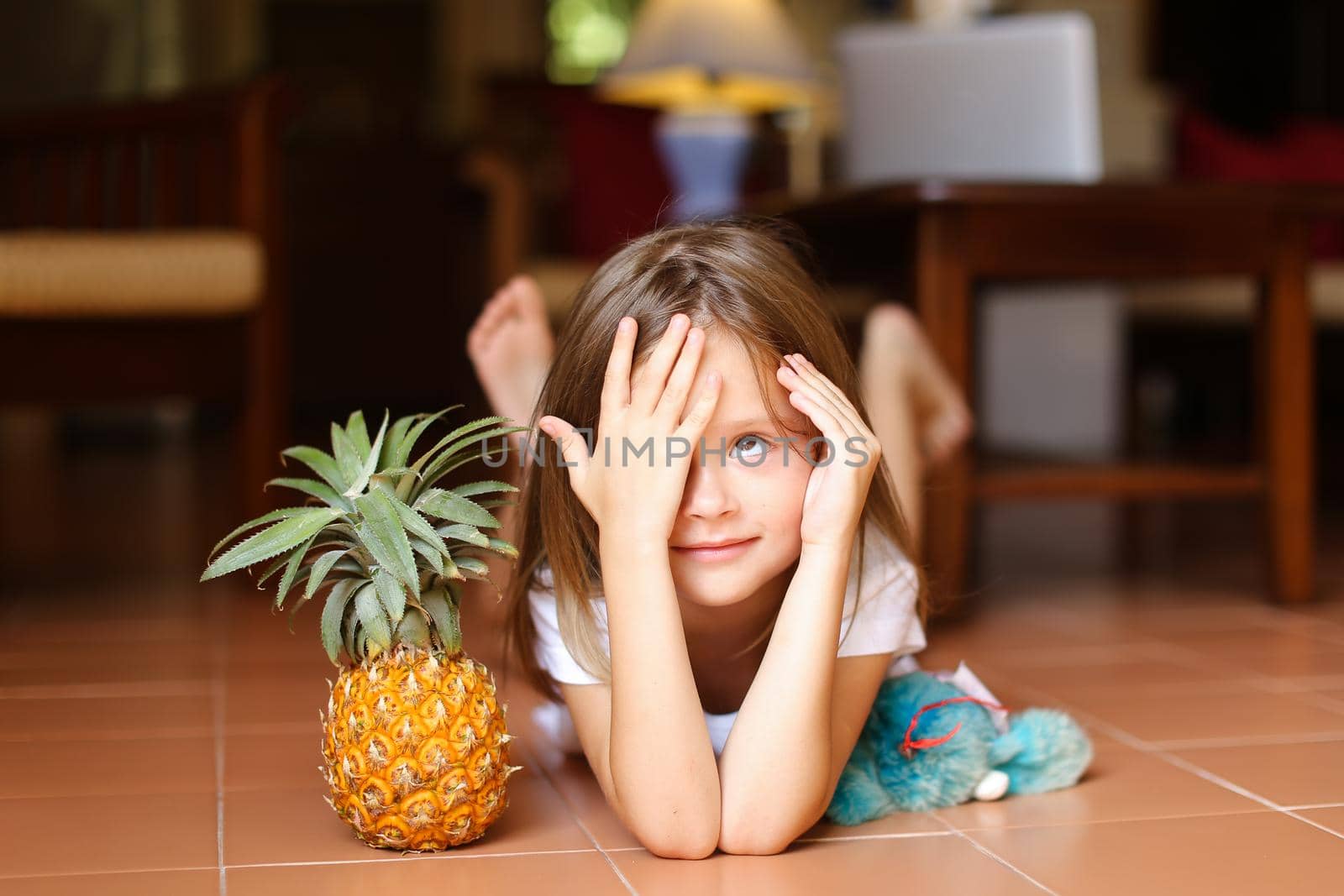 Portrait of little pretty girl lying on floor and playing with pineapple and toy, laptop in background. Concept of health vegeterian life and childhood.