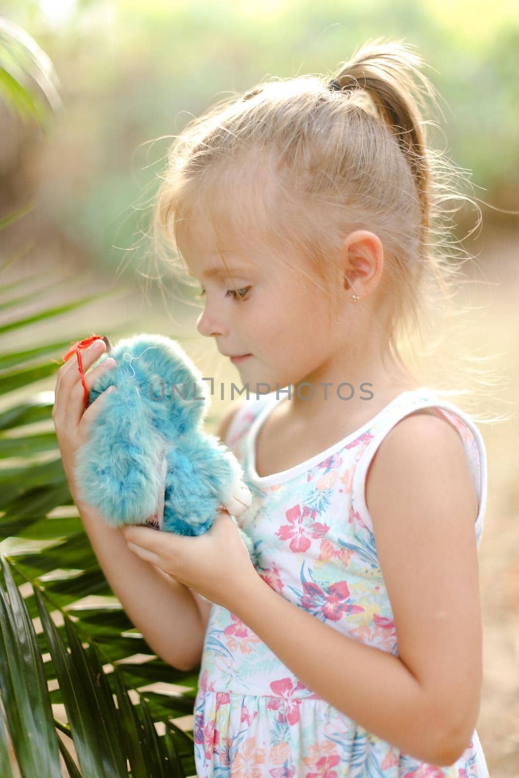 Portrait of little female kid playing with toy near plam leaf outside. Concept of tour with children to exotic countries, vacations with kids in Thailand.