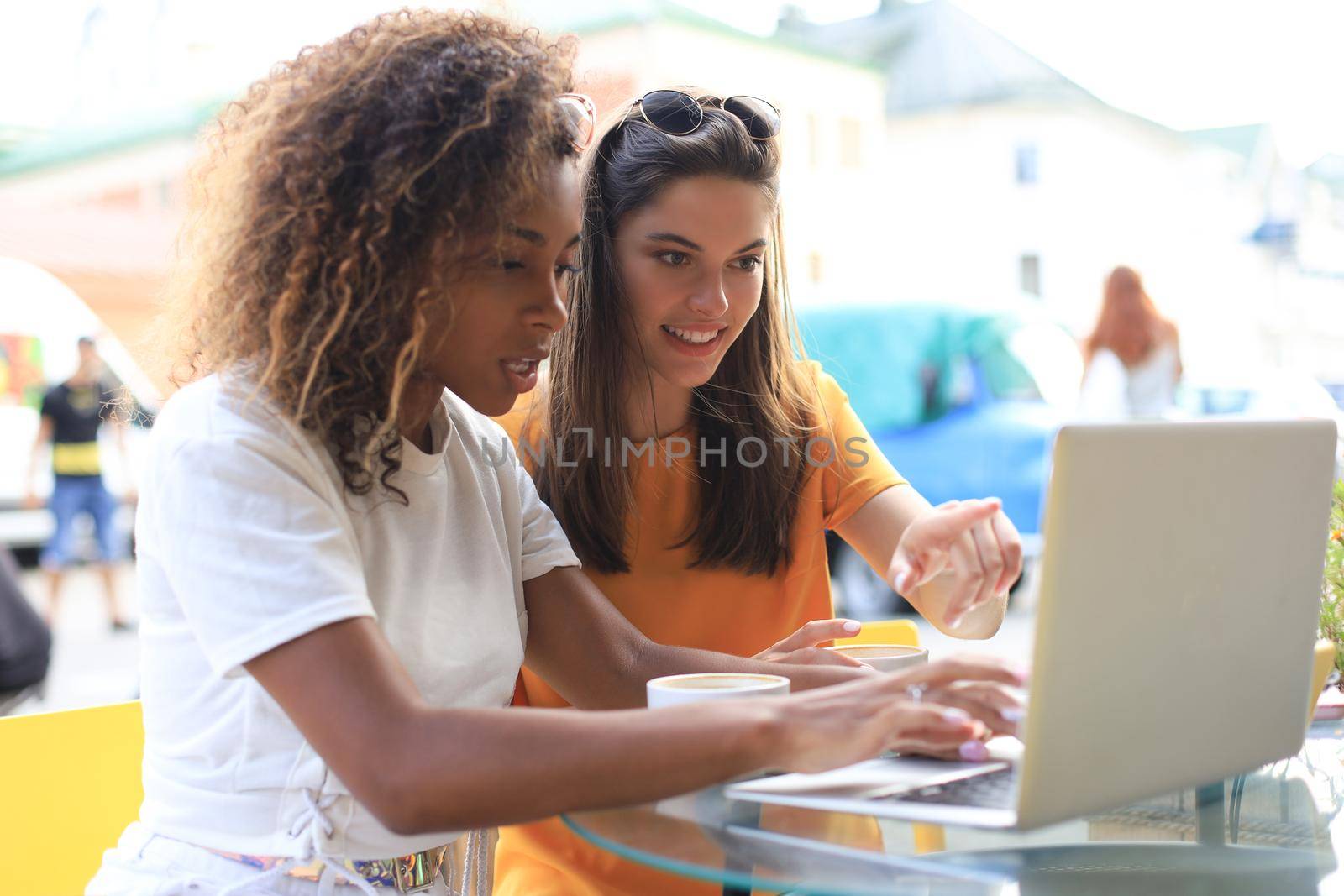 Two cheerful young girls friends sitting at cafe, having fun together, drinking coffee, using laptop