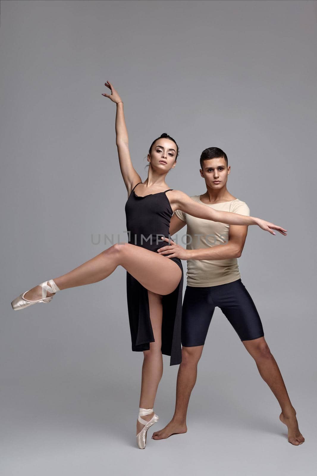 Pair of a graceful ballet dancers are posing over a gray studio background and looking at the camera. Handsome man in black shorts with beige t-shirt and beautiful woman in a black dress and white pointe shoes are dancing together. Ballet and contemporary choreography concept. Art photo.