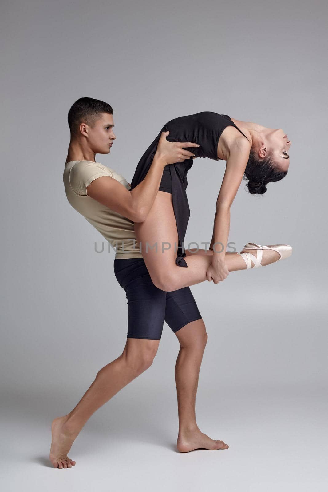 Pair of an adorable graceful ballet dancers are posing over a gray studio background. Handsome man in black shorts with beige t-shirt is holding a gorgeous woman in a black dress and white pointe shoes in his strong hands. Ballet and contemporary choreography concept. Art photo.