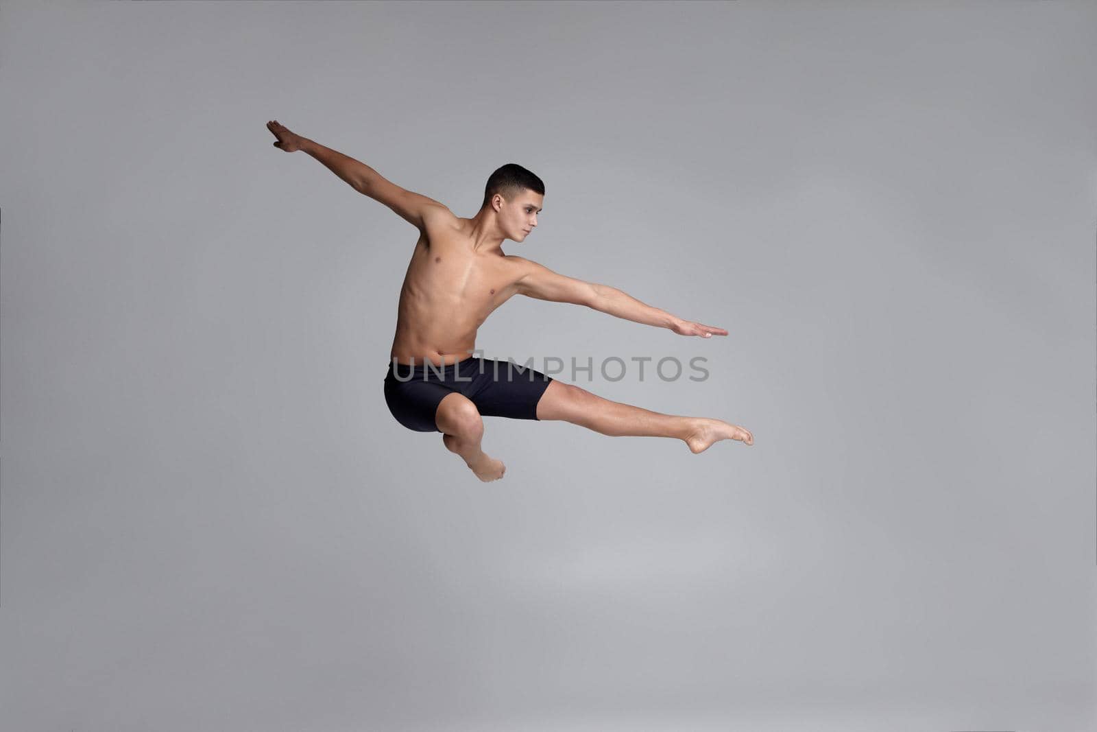 Photo of a handsome young male ballet dancer, dressed in a black shorts. He is jumping on a gray background in studio. Bare legs and torso. Ballet and contemporary choreography concept. Art photo.