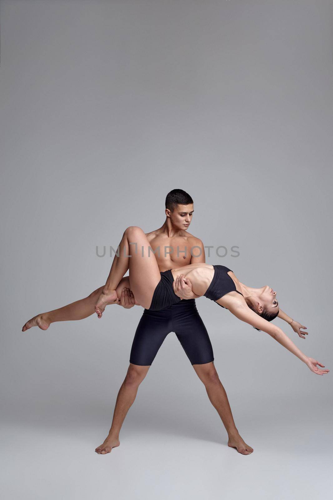 Two modern ballet dancers in black suits are posing over a gray studio background. Good-looking man in black shorts is holding a charming woman in a black swimwear. Ballet and contemporary choreography concept. Art photo.