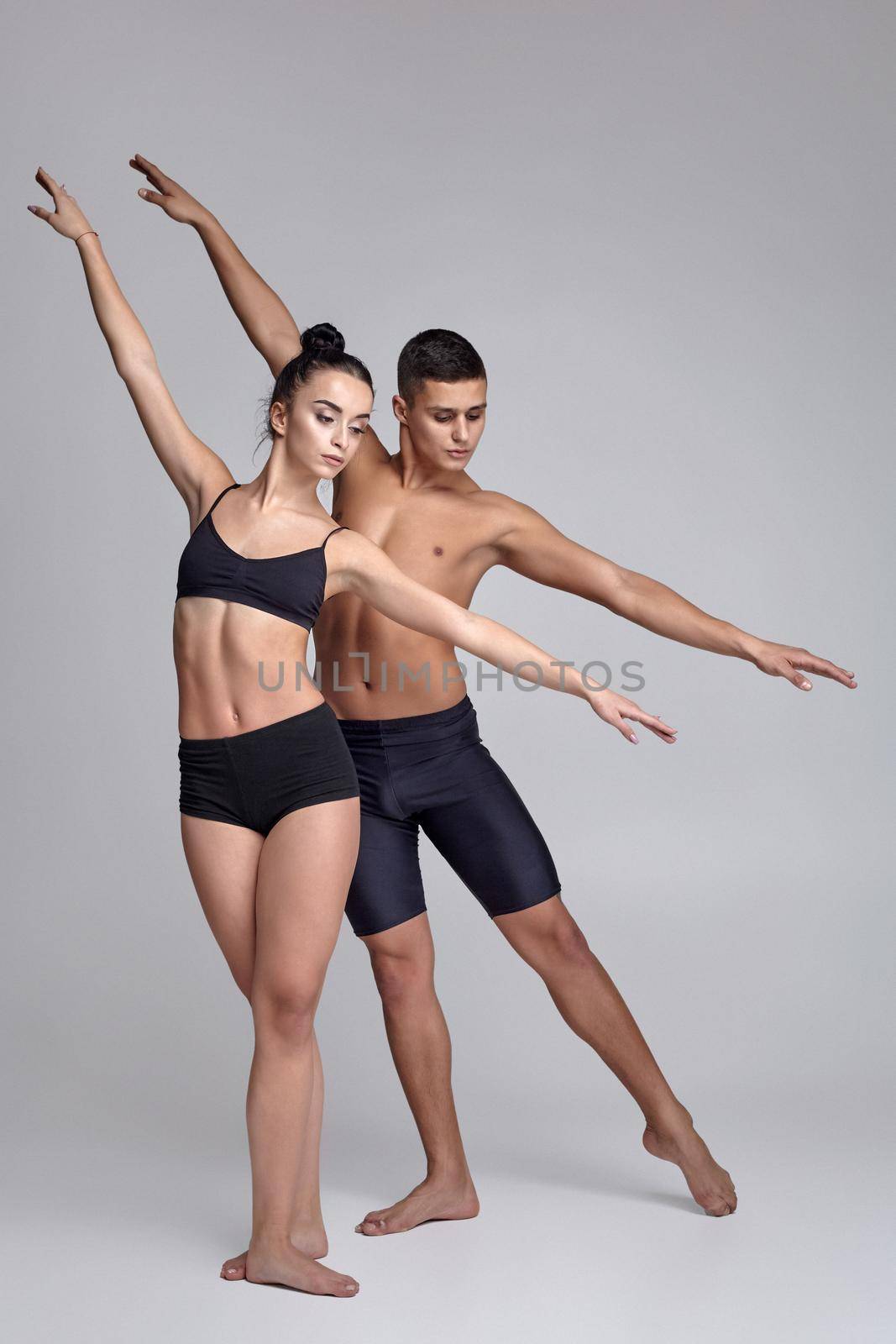 A pair of modern ballet dancers in black suits are posing over a gray studio background. Handsome man in black shorts and beautiful woman in a black swimwear are dancing together. Ballet and contemporary choreography concept. Art photo.