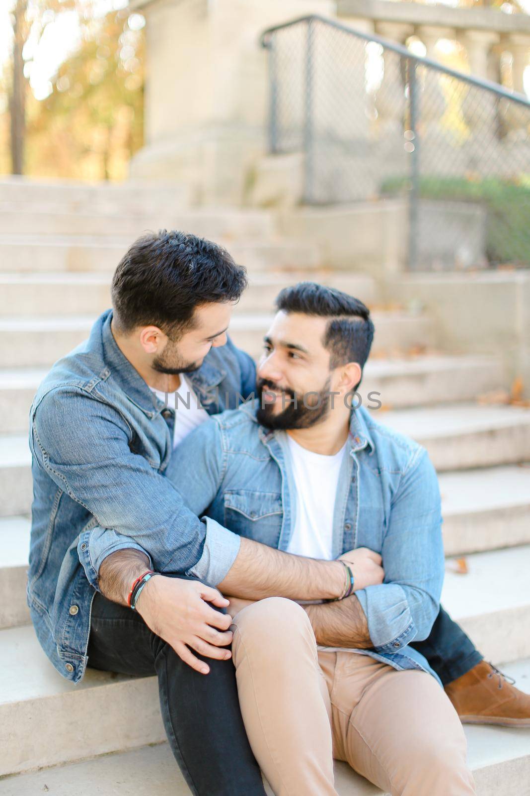 Caucasian european young gays sitting on concrete stairs and hugging, wearing jeans shirts. Concept of same sex couple and lgbt.