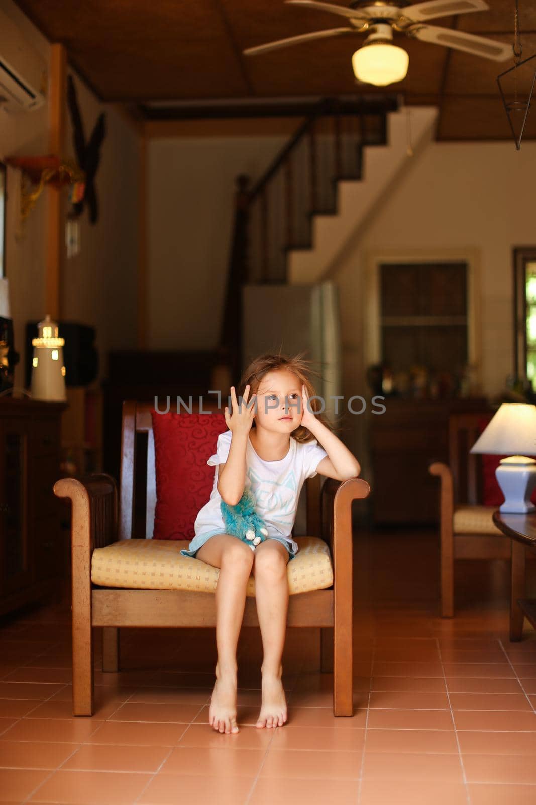 Little female child sitting in chair with in living room. Concept of leisure time for children and wooden interior.