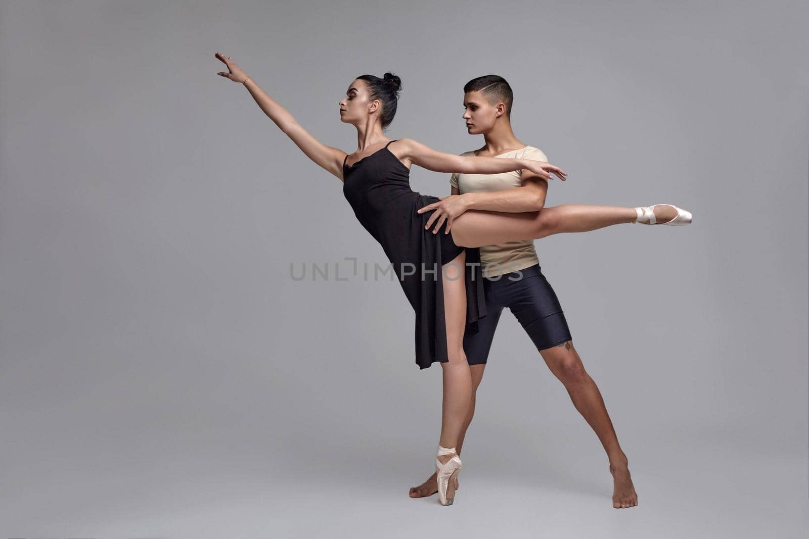 Pair of a graceful ballet dancers are posing over a gray studio background. Handsome man in black shorts with beige t-shirt and beautiful woman in a black dress and white pointe shoes are dancing together. Ballet and contemporary choreography concept. Art photo.