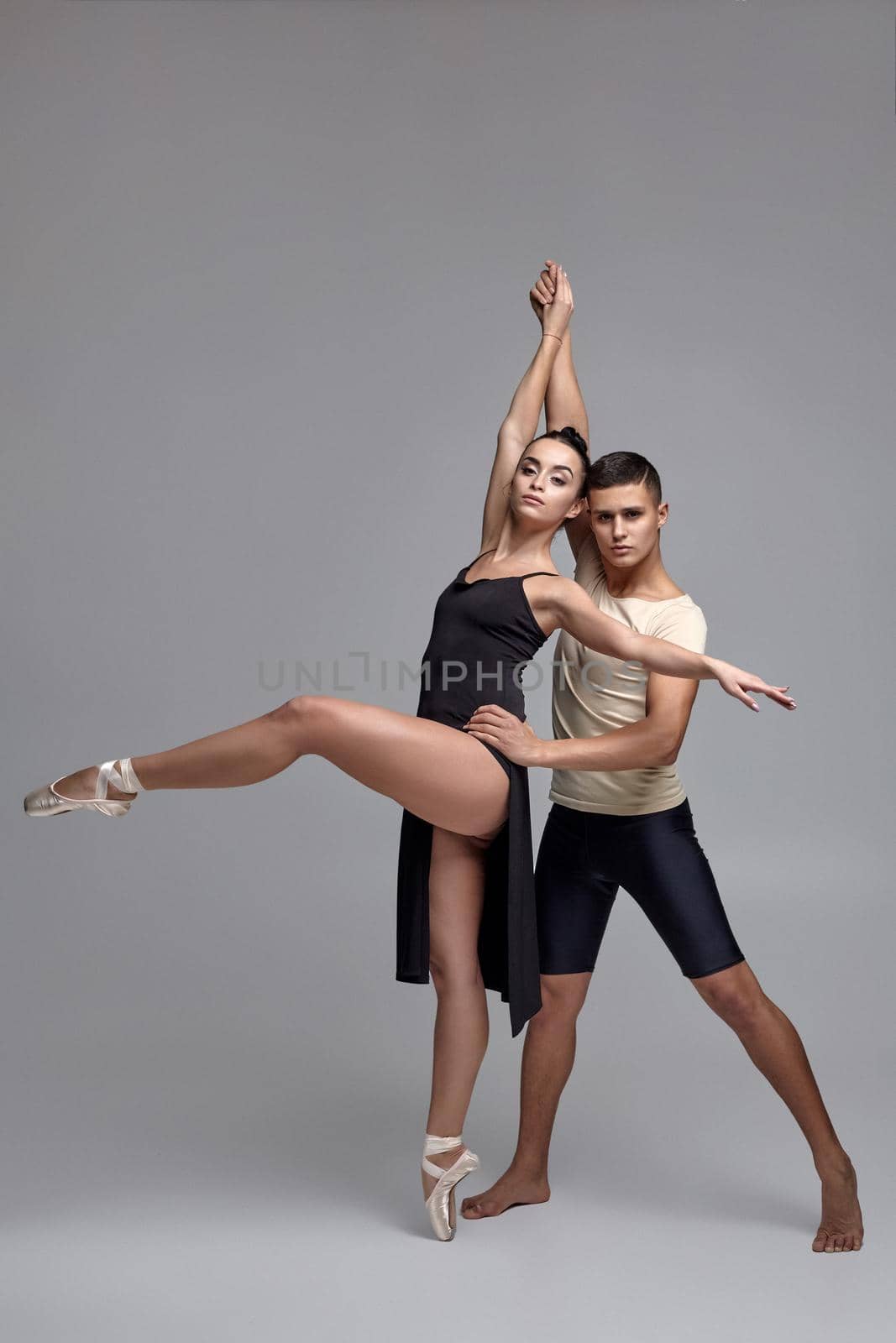 Pair of a modern ballet dancers are posing over a gray studio background and looking at the camera. Handsome man in black shorts with beige t-shirt and attractive woman in a black dress and white pointe shoes are dancing together. Ballet and contemporary choreography concept. Art photo.