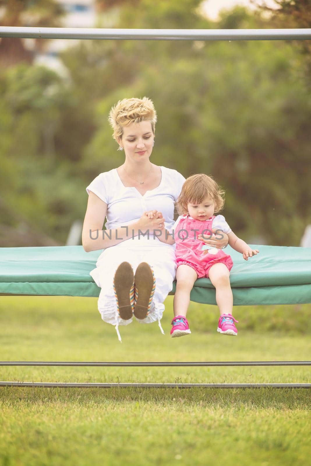 A beautiful young mother and her happy little daughter enjoying free time while swinging on bench at backyard