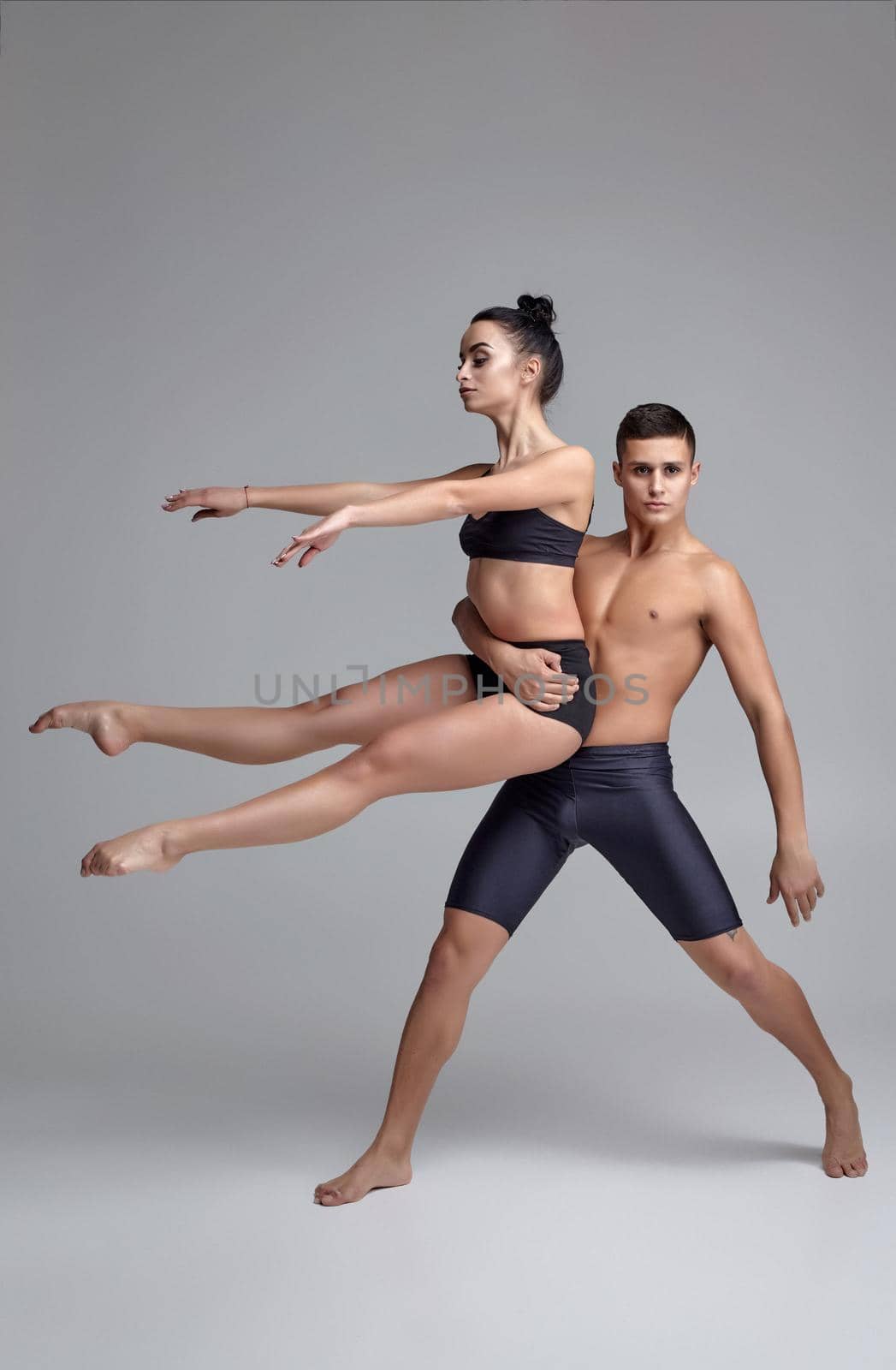 A couple of a ballet dancers in black suits are posing over a gray studio background. Handsome male in black shorts is holding in his hands a beautiful female in a black swimwear. Ballet and contemporary choreography concept. Art photo.