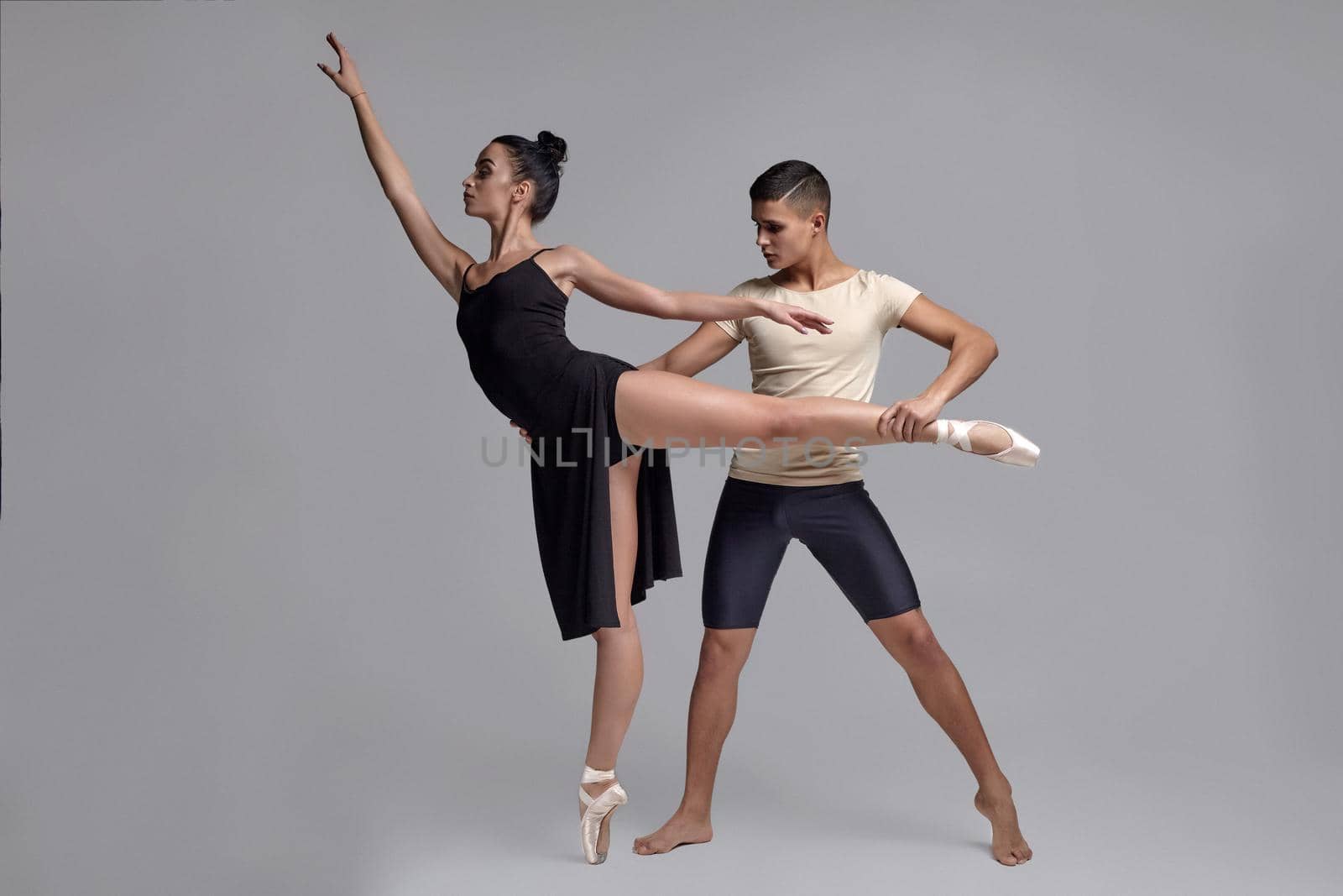 Couple of a young graceful ballet dancers are posing over a gray studio background. Handsome male in black shorts with beige t-shirt and beautiful female in a black dress and white pointe shoes are dancing together. Ballet and contemporary choreography concept. Art photo.