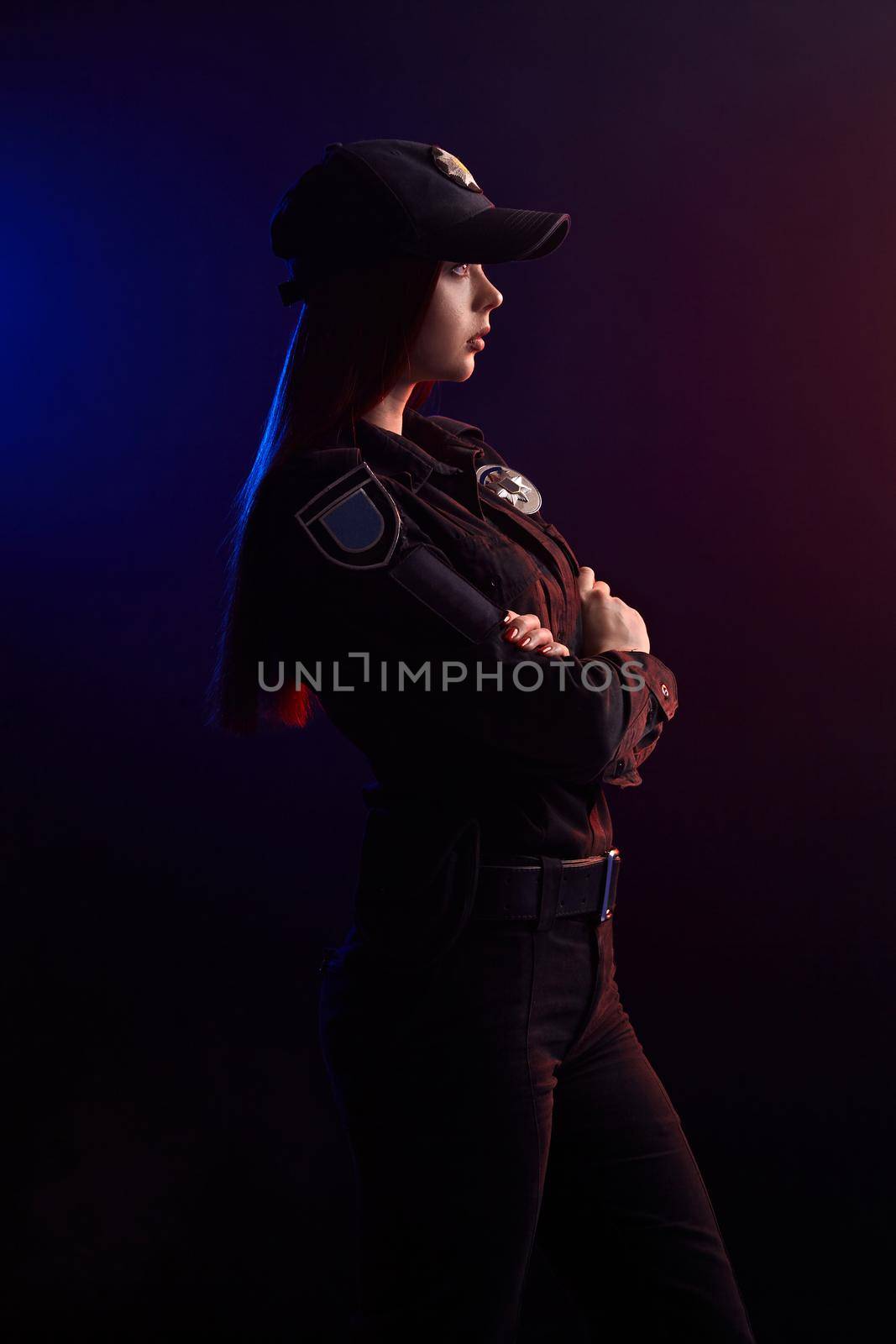 Pretty redheaded female police officer in a uniform and a cap is posing sideways with crossed hands and looking away, against a black background with red and blue backlighting.