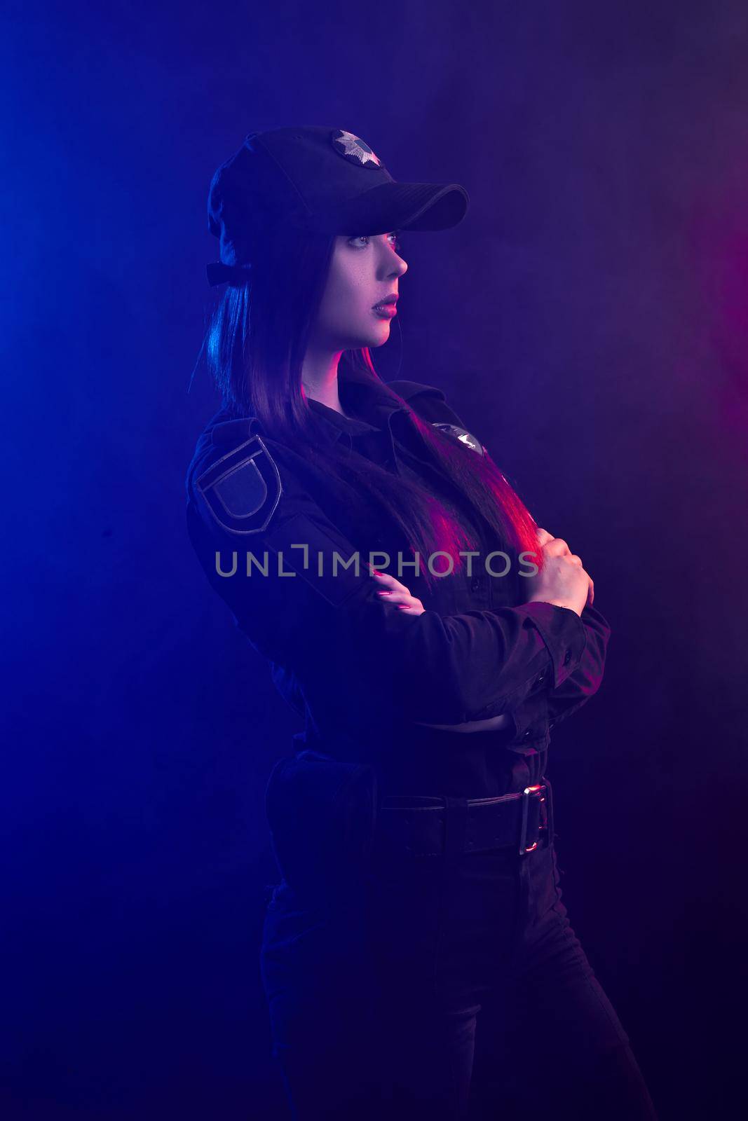 Alluring redheaded woman police officer in a uniform and a cap, with bright make-up, is standing sideways and looking away against a black background with red and blue backlighting.