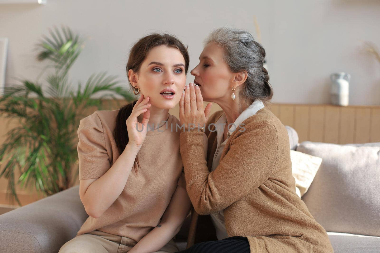 Happy adult daughter whispering secret to her middle mother at home, gossiping, sharing secret. Beautiful woman hearing good news from mature mum, trusted relations.