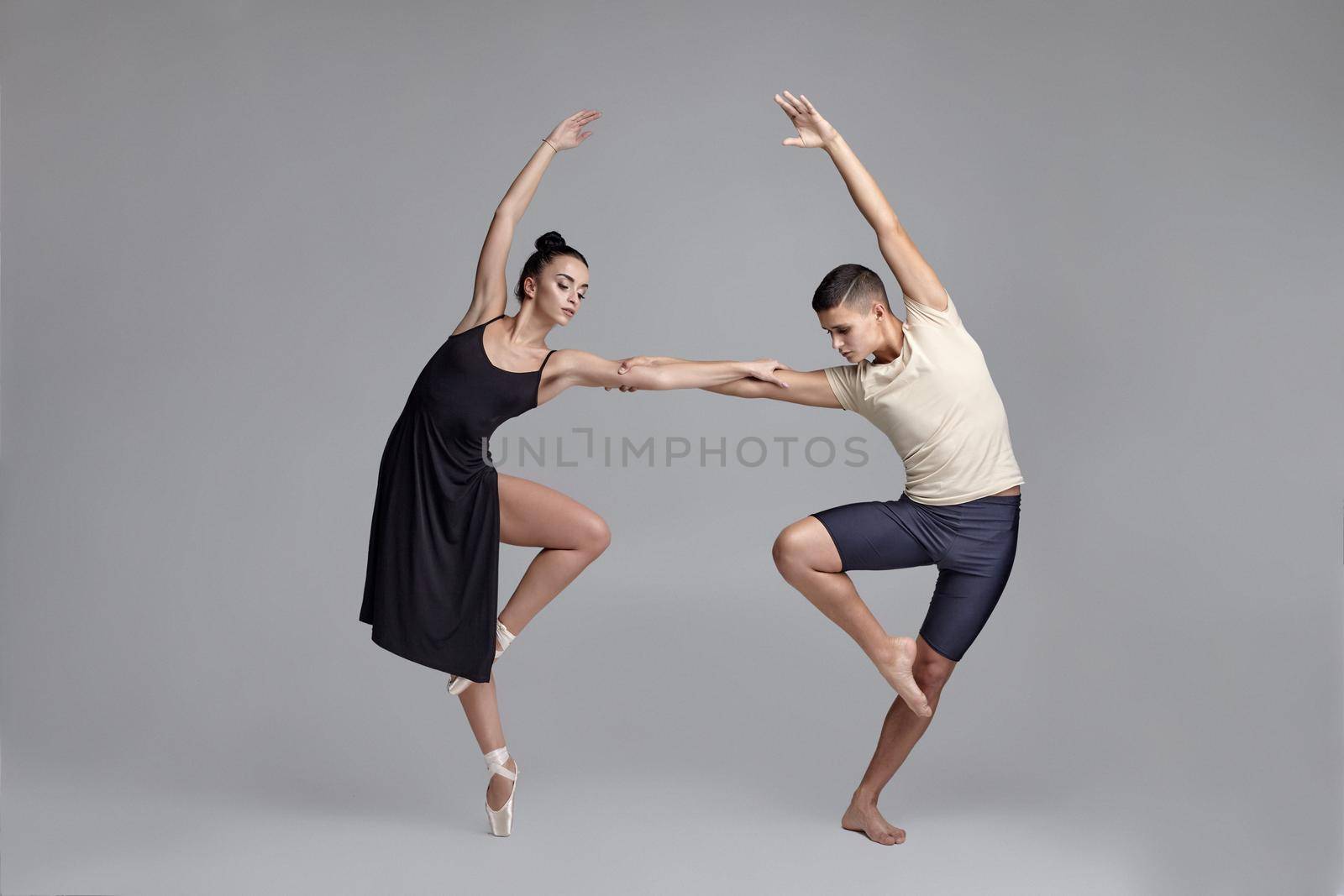 Photo of a young athletic ballet dancers posing over a gray studio background. Handsome male in black shorts with beige t-shirt and pretty girl in a black dress and white pointe shoes are dancing together. Ballet and contemporary choreography concept. Art photo.