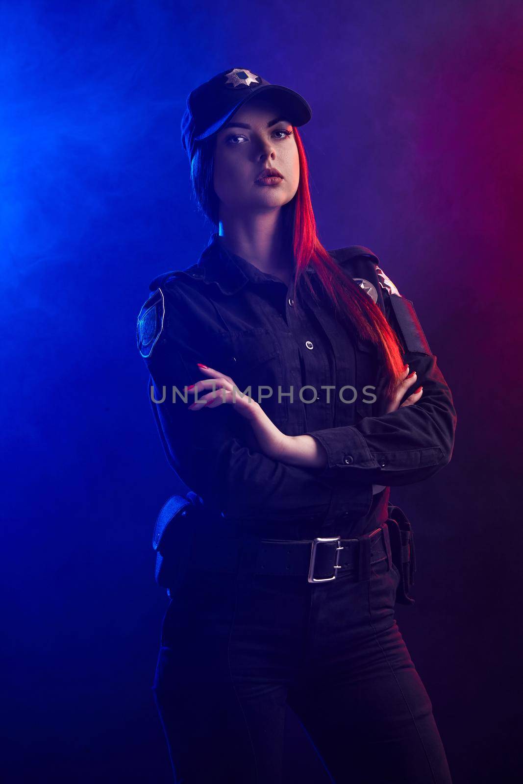 Wonderful redheaded female police officer in a uniform and a cap is posing with crossed hands and looking at the camera against a black background with red and blue backlighting. Defender of citizens is ready to enforce a law and stop a crime.