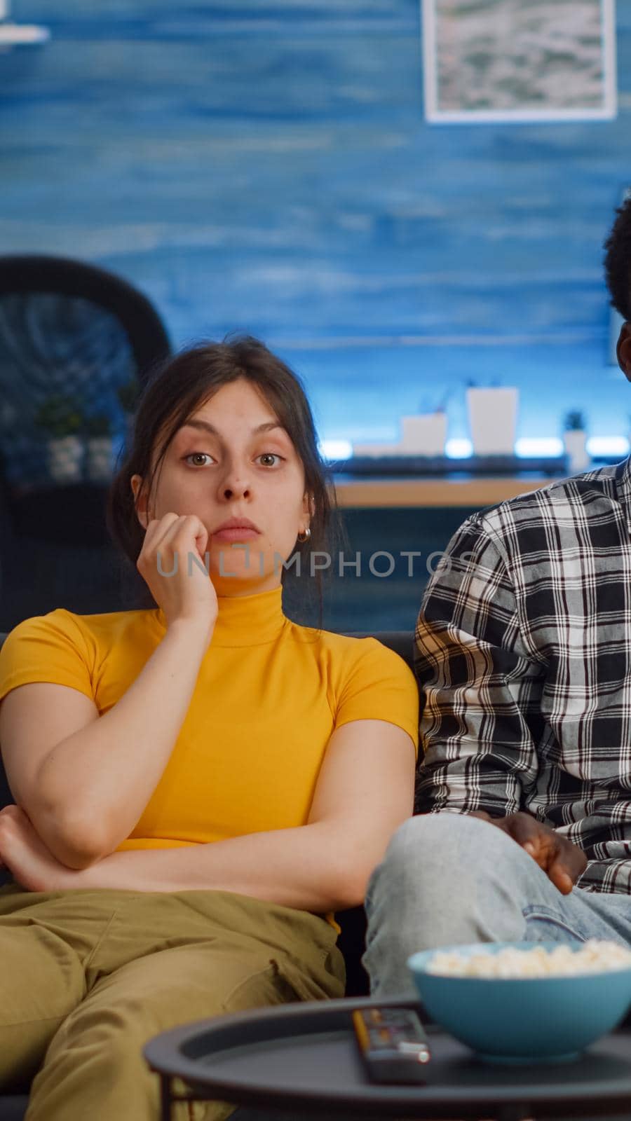 Young interracial couple watching television eating popcorn in living room. Married multi ethnic people looking at camera and having snack enjoying relaxing activity. Mixed race lovers