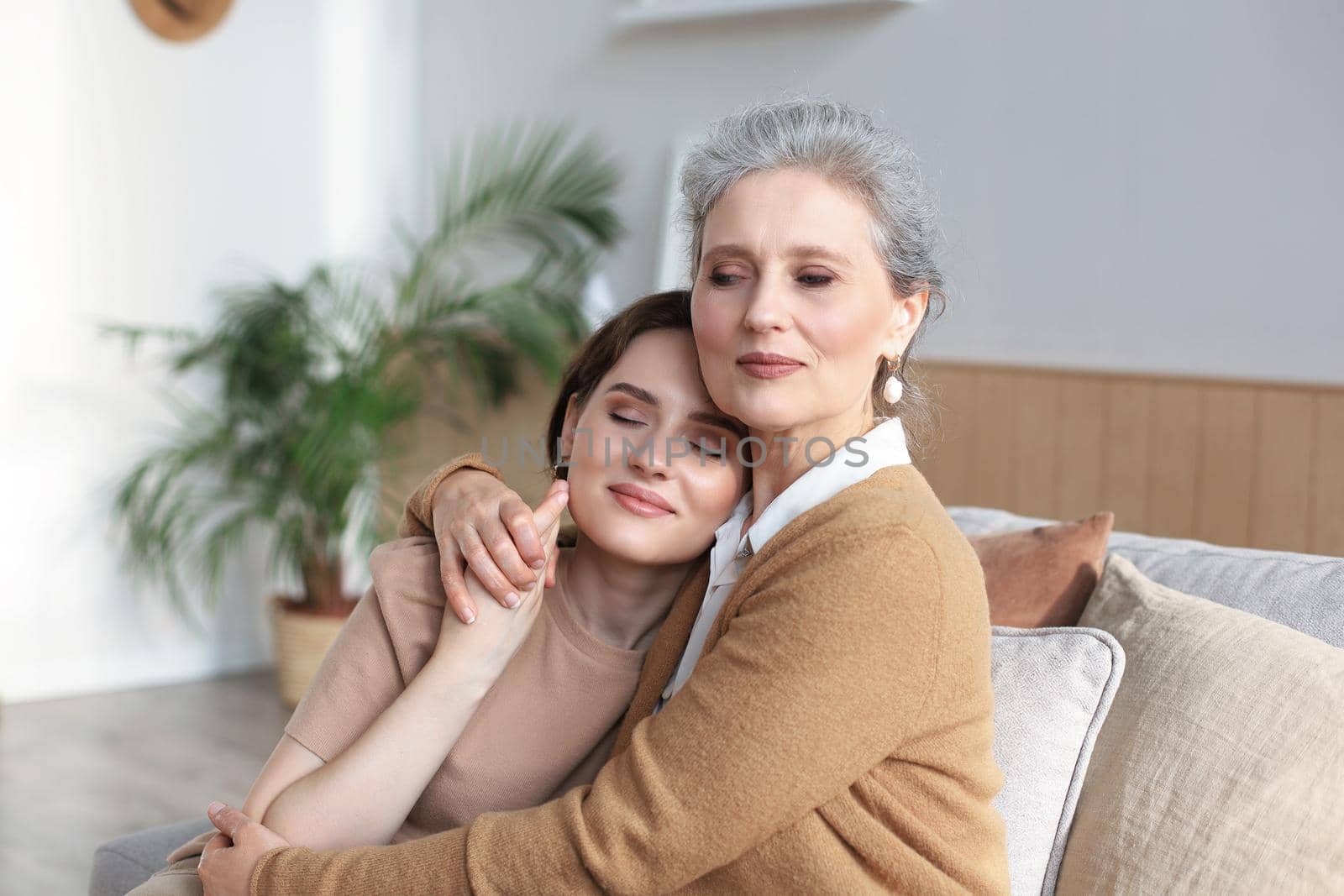Cheerful young woman is embracing her middle aged mother with closed eyes hugging, touching cheeks, sitting on couch at home. Happy trusted relations. Family concept. by tsyhun