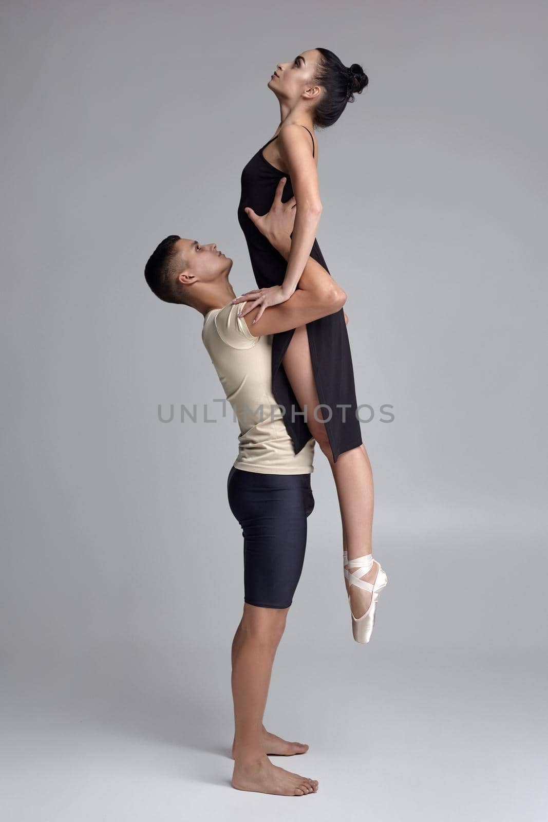 Pair of a graceful ballet dancers are posing over a gray studio background. Handsome man in black shorts with beige t-shirt is holding a beautiful woman in a black dress and white pointe shoes in his strong hands. Ballet and contemporary choreography concept. Art photo.