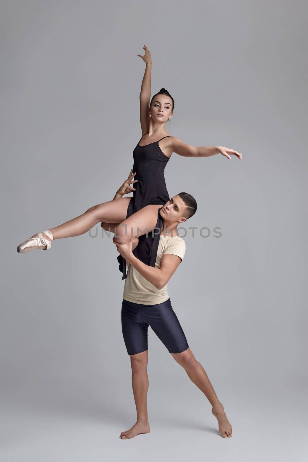 Studio shot of a modern ballet dancers posing over a gray background. Strong man in black shorts with beige t-shirt is holding a beautiful girl in a black dress and white pointe shoes in his strong hands. Ballet and contemporary choreography concept. Art photo.
