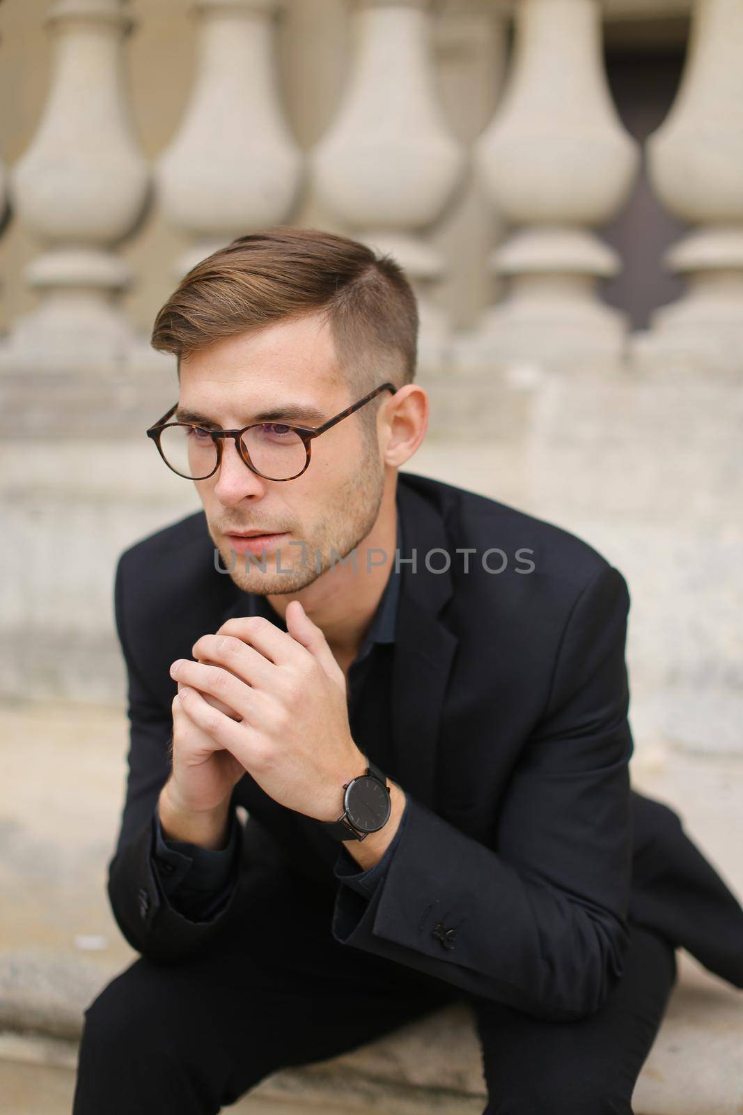 Young fashionable man sitting on sidewalk and leaning on concrete railing of building, wearing black suit and glasses. Concept of walkig in city, urban photo session and male person model.