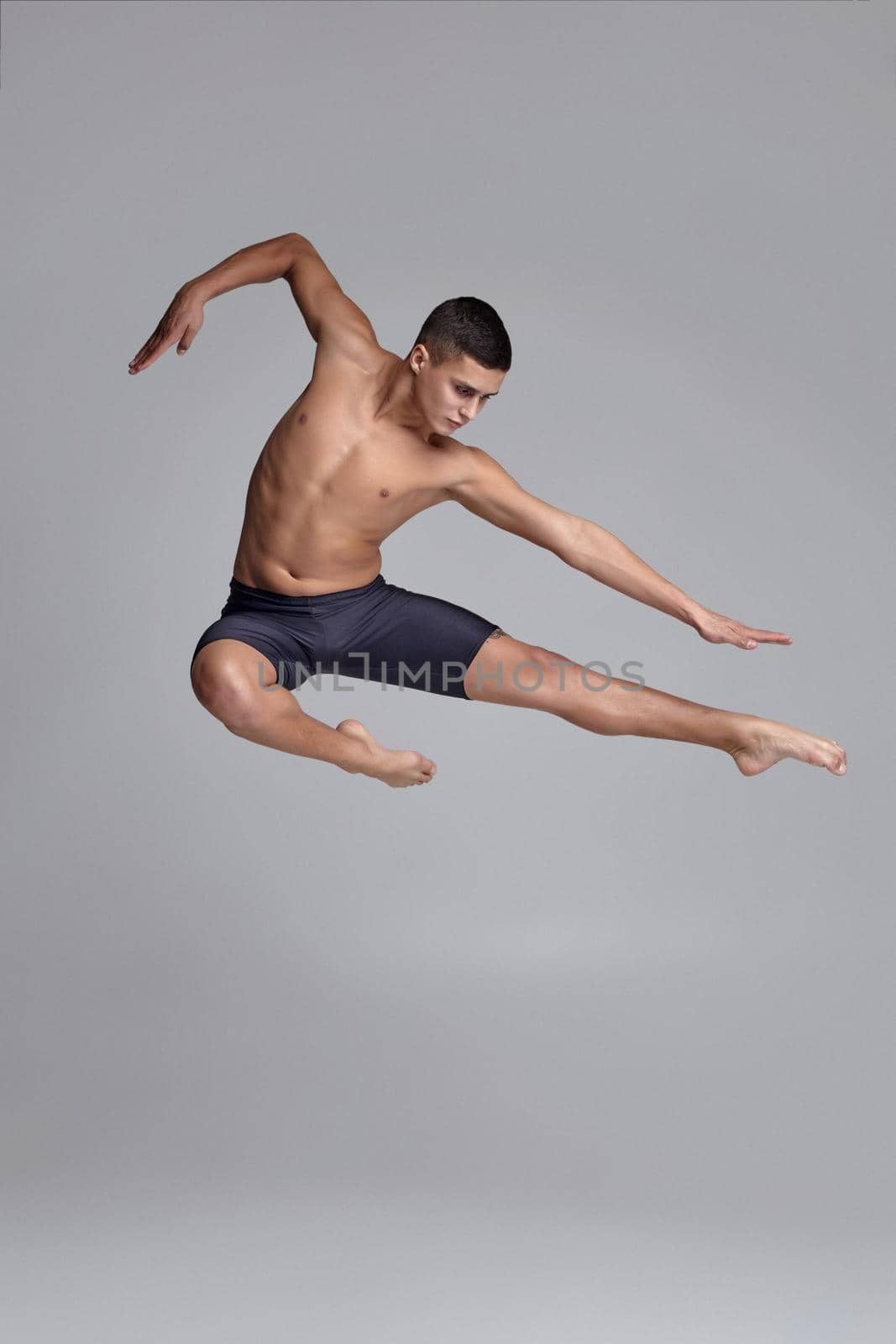 Portrait of a stately man ballet dancer, dressed in a black shorts. He is jumping on a gray background in studio. Bare legs and torso. Ballet and contemporary choreography concept. Art photo.