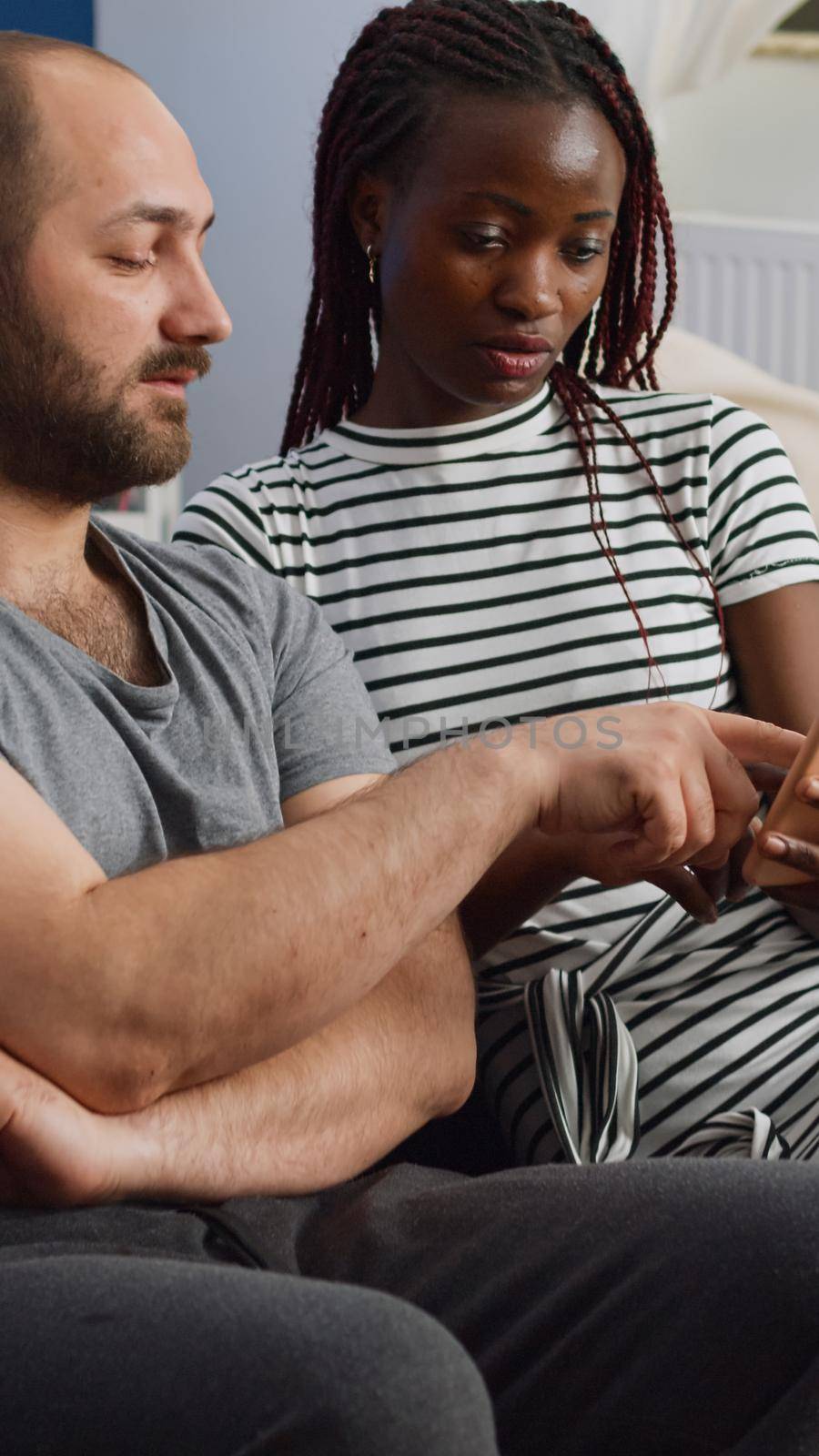 Young interracial couple looking at smartphone while black woman holding device at home. Mixed race husband and wife using technology and talking while sitting on couch in living room