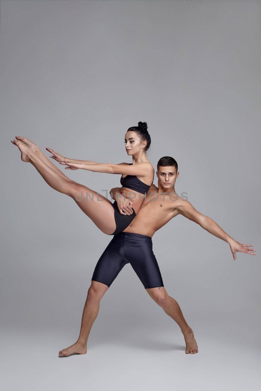 A couple of a young ballet dancers in black suits are posing over a gray studio background. Attractive man in black shorts is holding in his hands a beautiful woman in a black swimwear. Ballet and contemporary choreography concept. Art photo.