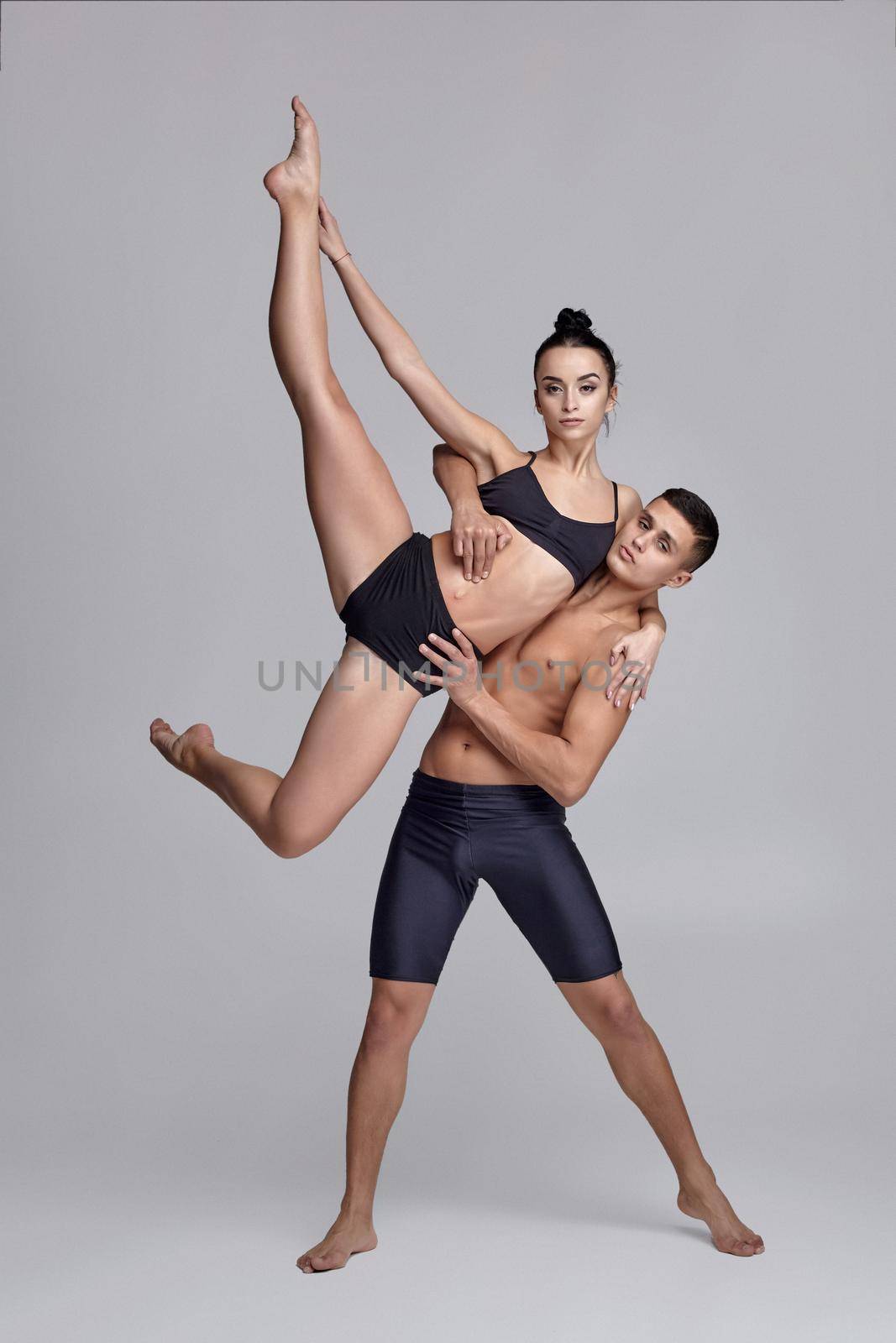 A couple of a strong ballet dancers in black suits are posing over a gray studio background. Handsome man in black shorts is holding in his hands a pretty woman in a black swimwear. Ballet and contemporary choreography concept. Art photo.
