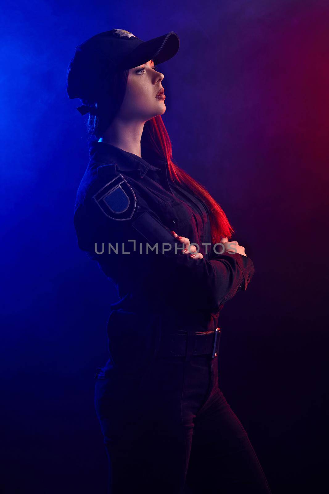 Serious female police officer is posing for the camera against a black background with red and blue backlighting. by nazarovsergey
