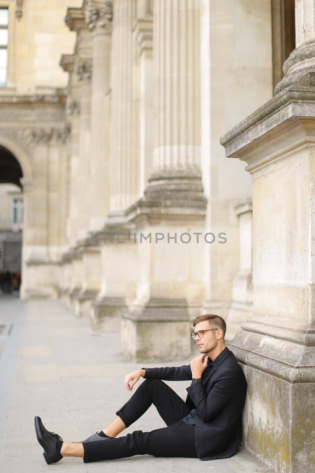 Young caucasian man sitting on sidewalk ground and wearing black suit. by sisterspro