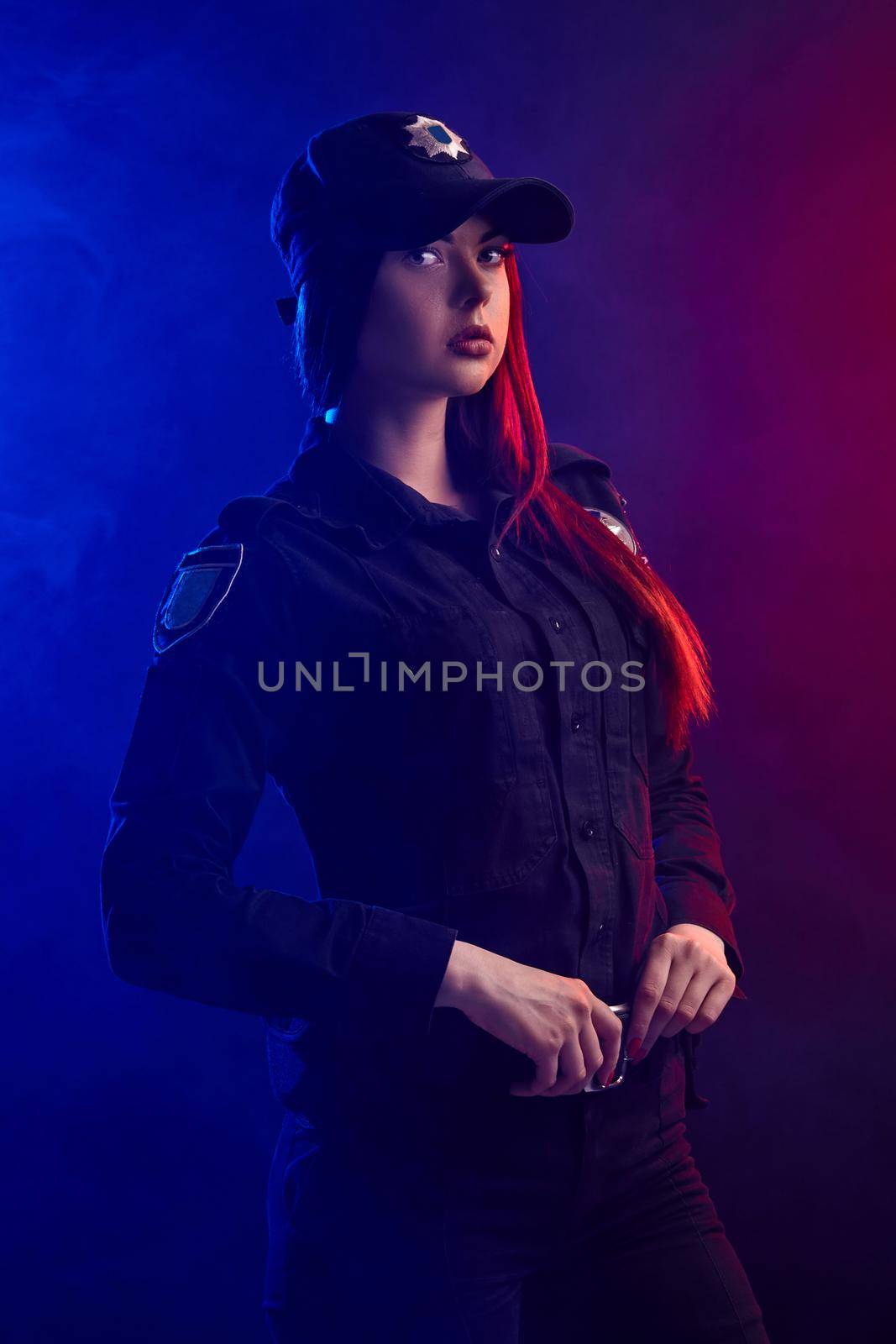 Cute redheaded woman police officer in a uniform and a cap, with bright make-up, is standing sideways, holding her belt and looking at the camera against a black background with red and blue backlighting.