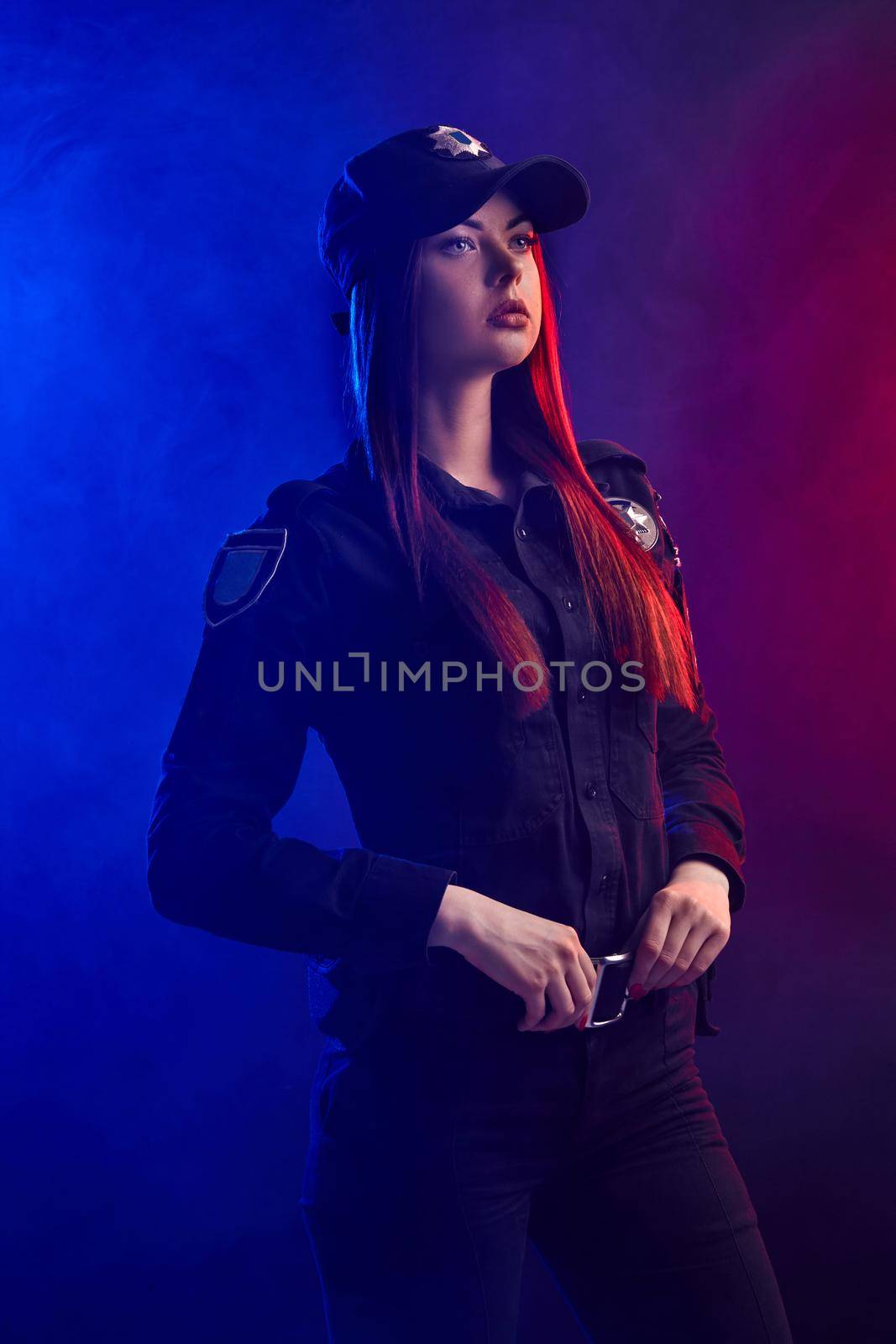 Attractive ginger girl police officer in a uniform and a cap, with bright make-up, is standing sideways, holding her belt and looking away against a black background with red and blue backlighting.
