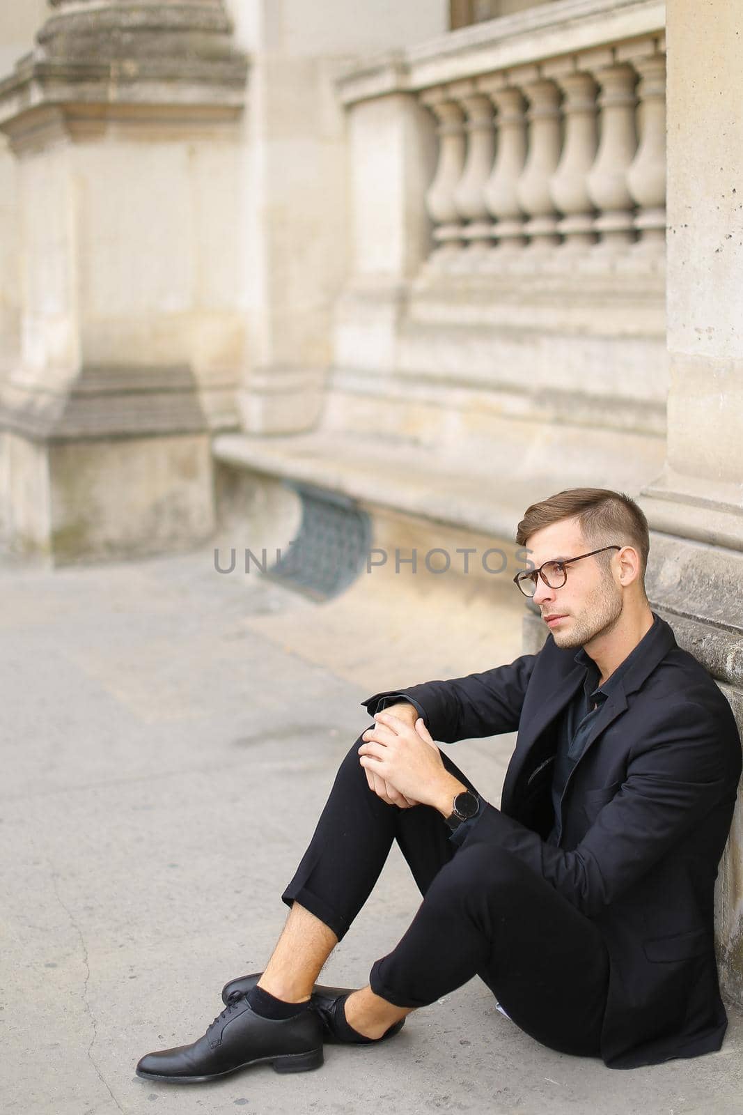 Young stylish man sitting on sidewalk ground and wearing black suit, leaning on concrete banister. Concept of walking in cty and male fashionable model.