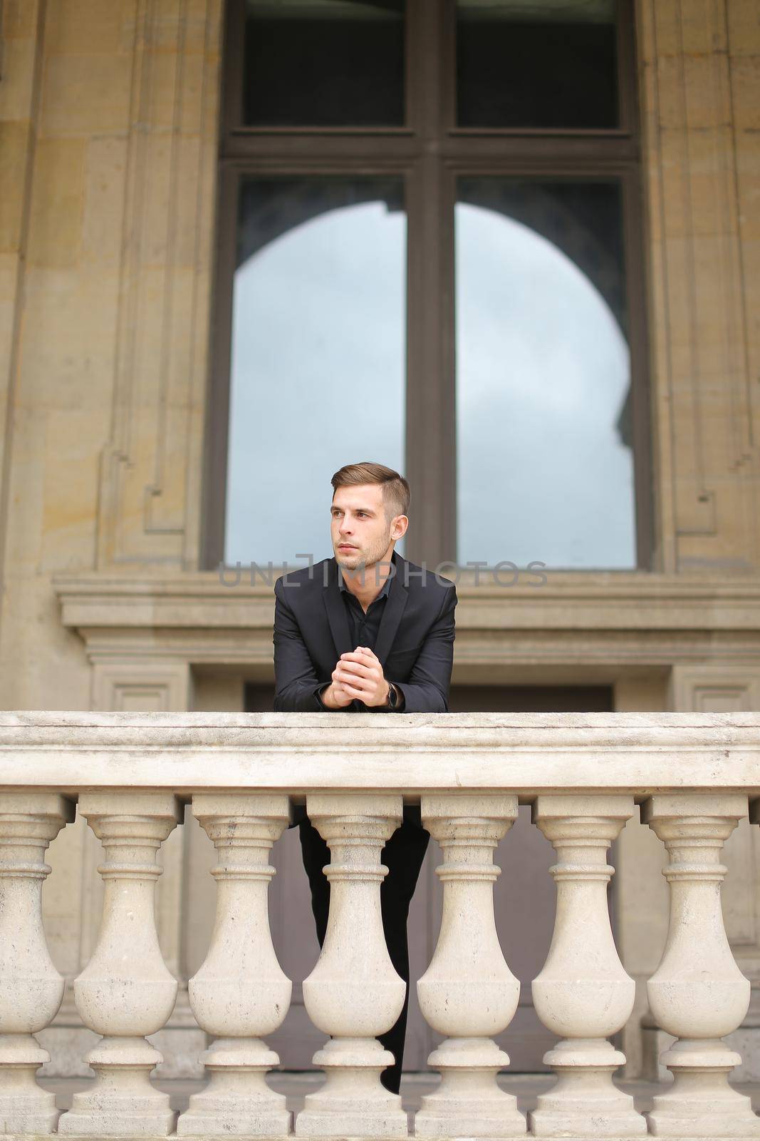 Young european boy leaning on balcony concrete railing in Paris. Concept of male fashon moden and architecture. Guy wearing black suit.