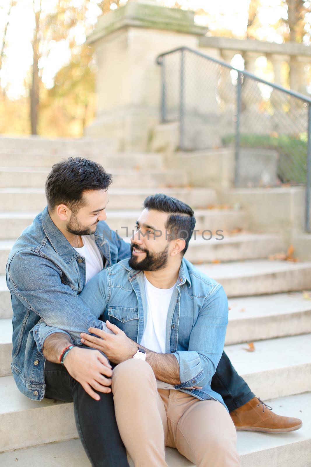 Caucasian american young gays sitting on concrete stairs and hugging, wearing jeans shirts. Concept of same sex couple and lgbt.