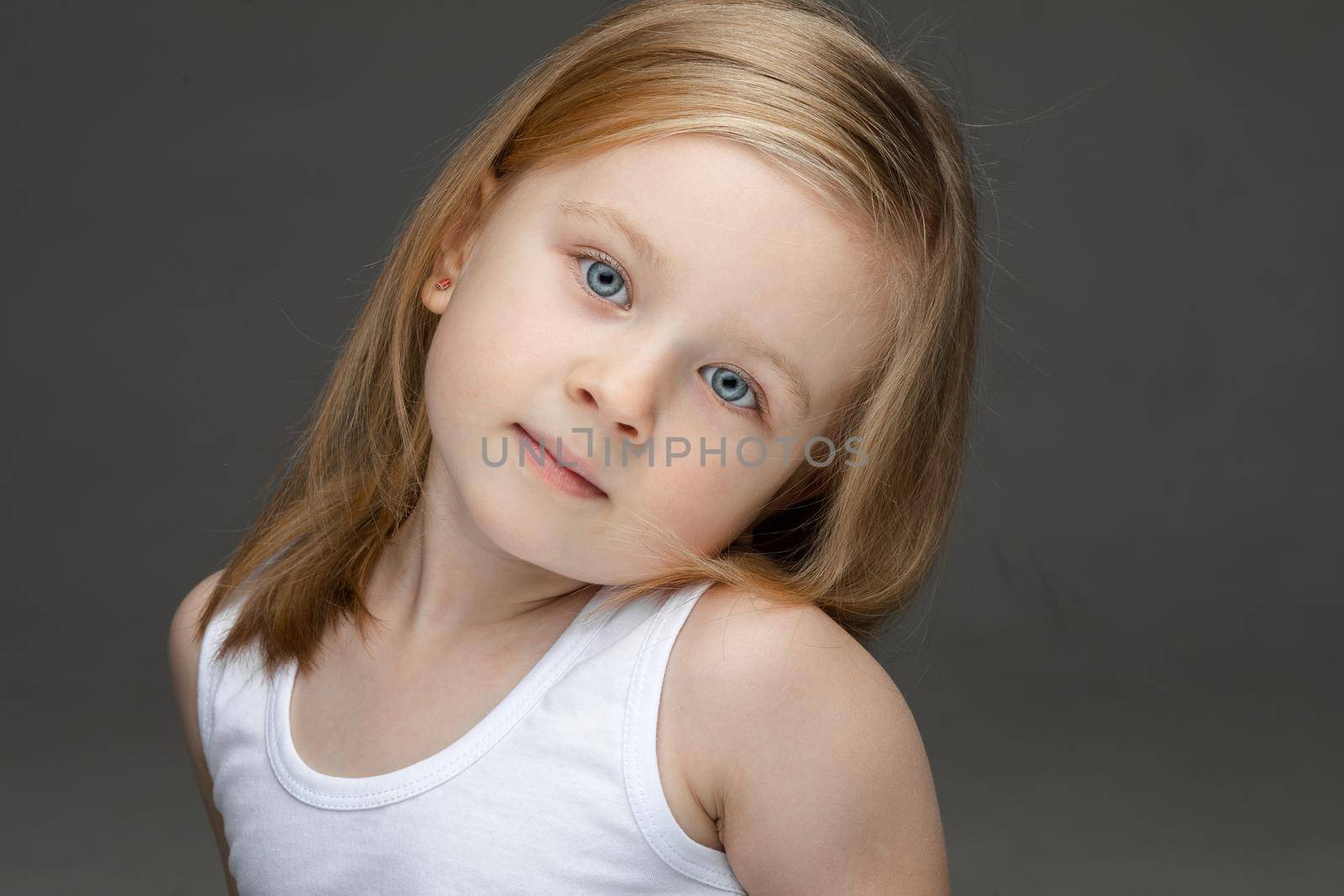 Calm cute little girl wearing a white undershirt looking at the camera while standing on grey background. Childhood concept