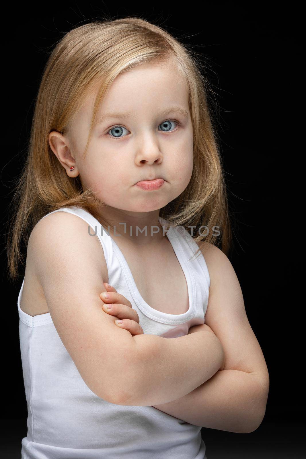 Upset child in a white undershirt standing on black background and feeling bored. Childhood concept