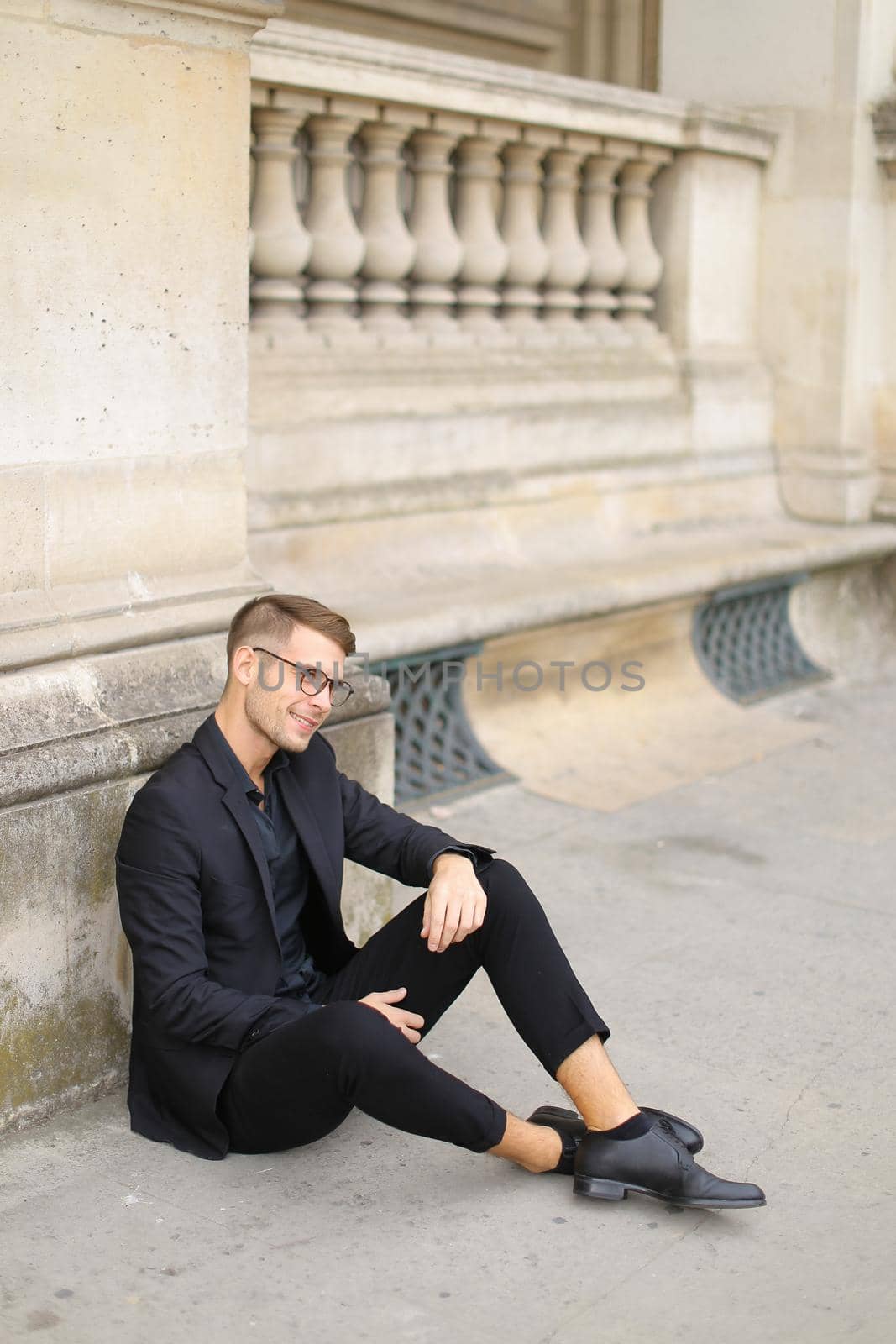 Young handsome man sitting on sidewalk ground and wearing black suit, leaning on concrete banister. Concept of walking in cty and male fashionable model.