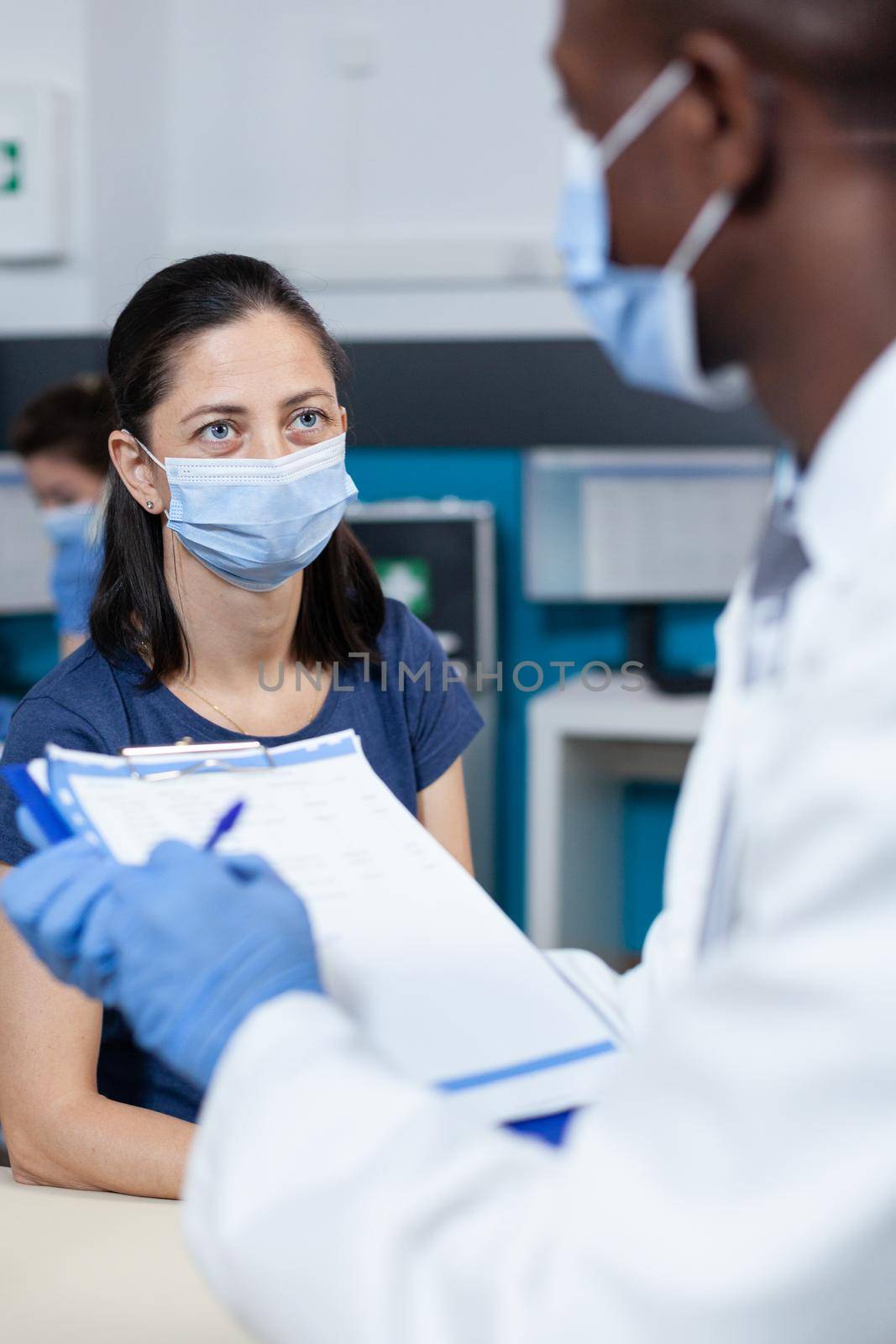 Closeup of african american pediatrician doctor writing ill diagnostic on clipboard explaining healthcare treatment to parent during clinical appointment in hospital office. Therapist with face mask