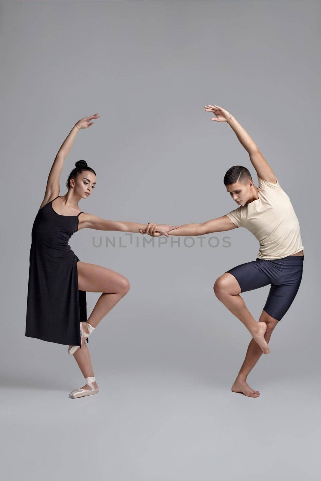 Two young ballet dancers are posing over a gray studio background. Attractive man in black shorts with beige t-shirt and beautiful woman in a black dress and white pointe shoes are dancing together. Ballet and contemporary choreography concept. Art photo.