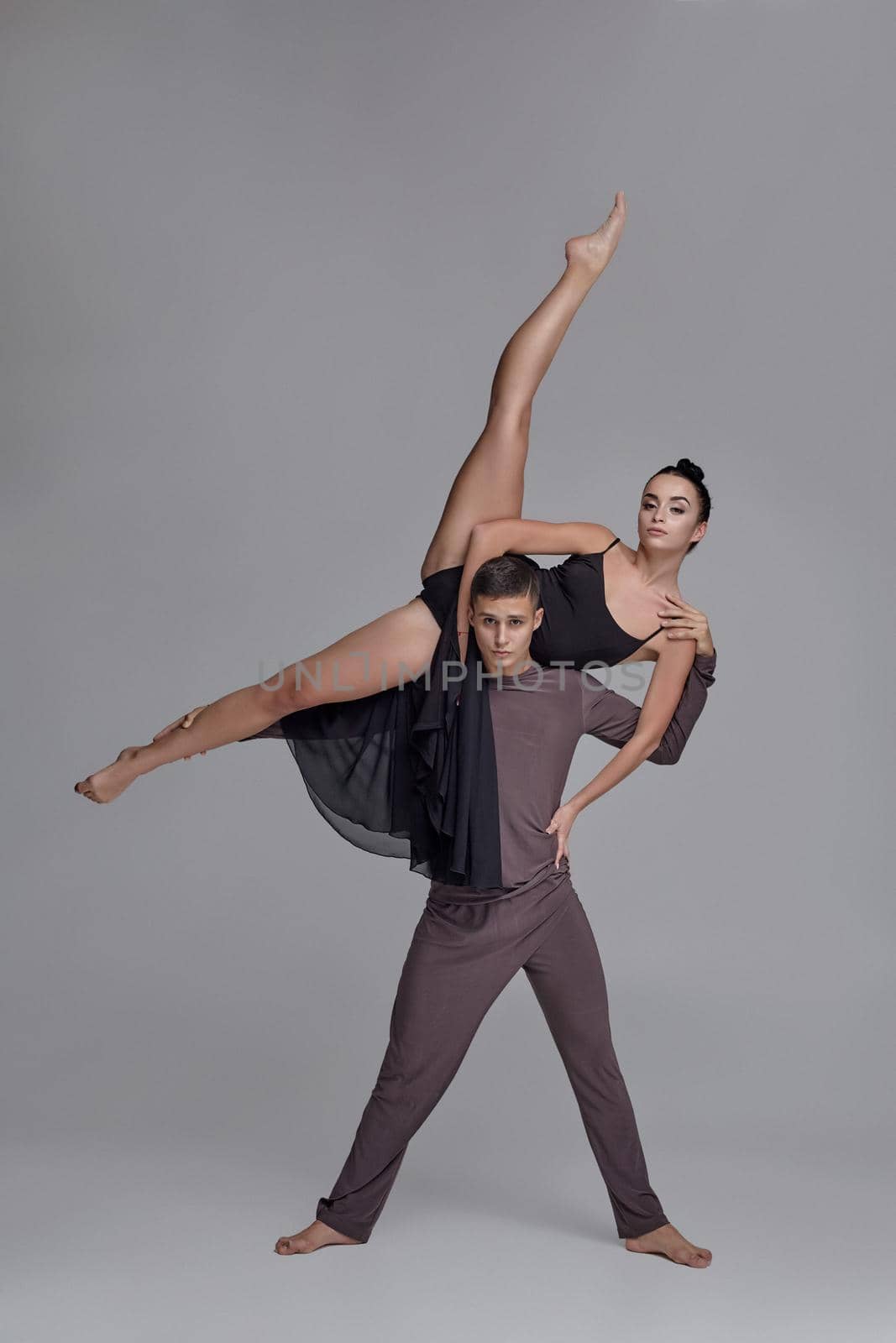 Pair of a graceful ballet dancers are posing over a gray studio background. Handsome man in a gray tracksuit and beautiful woman in a black dress are dancing together. Ballet and contemporary choreography concept. Art photo.
