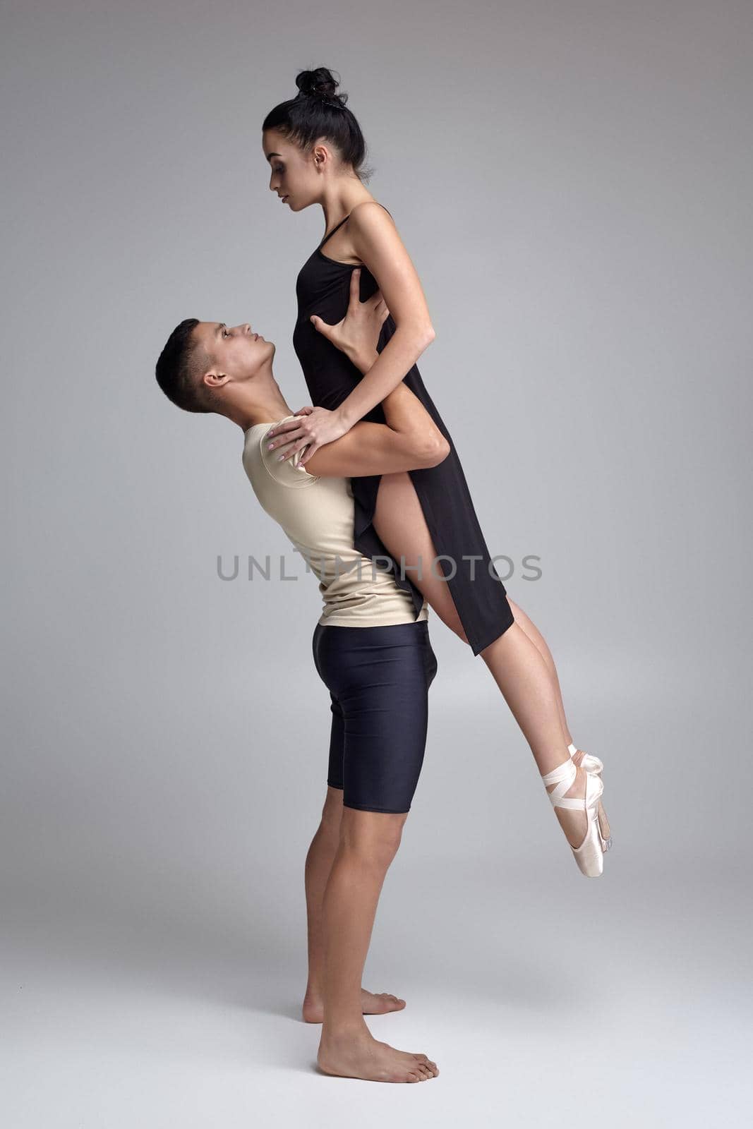 Couple of a young ballet dancers are posing over a gray studio background. Handsome man in black shorts with beige t-shirt is holding a beautiful woman in a black dress and white pointe shoes in his strong hands. Ballet and contemporary choreography concept. Art photo.