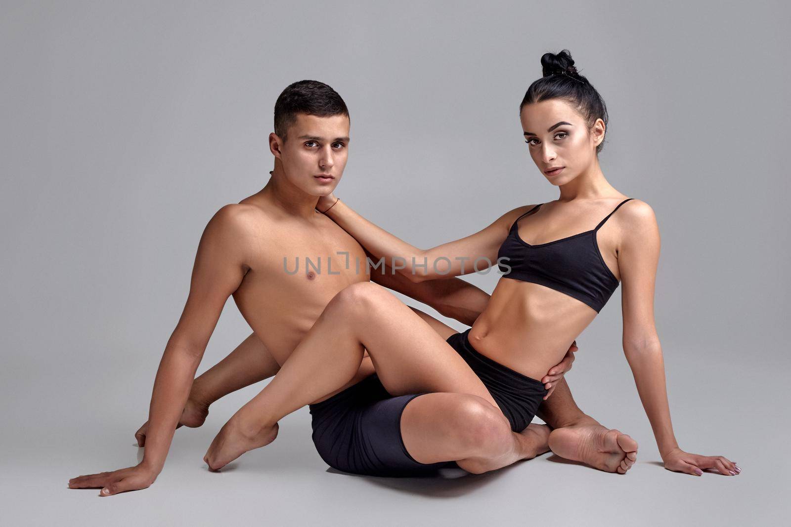 A pair of adult ballet dancers in black suits are posing over a gray studio background. Handsome man in black shorts and beautiful woman in a black swimwear are embracing each other and looking at the camera. Contemporary choreography concept. Art photo.