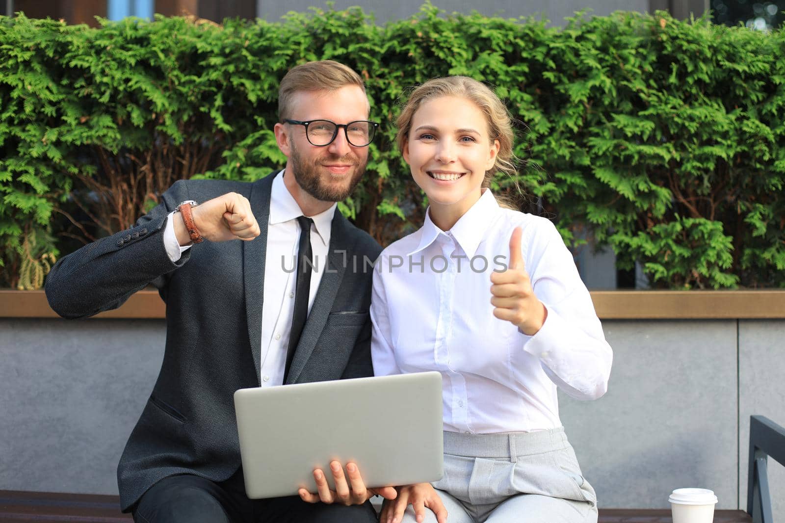 Office colleagues using laptop computer and showing thumbs up while sitting on a bench outdoor. by tsyhun