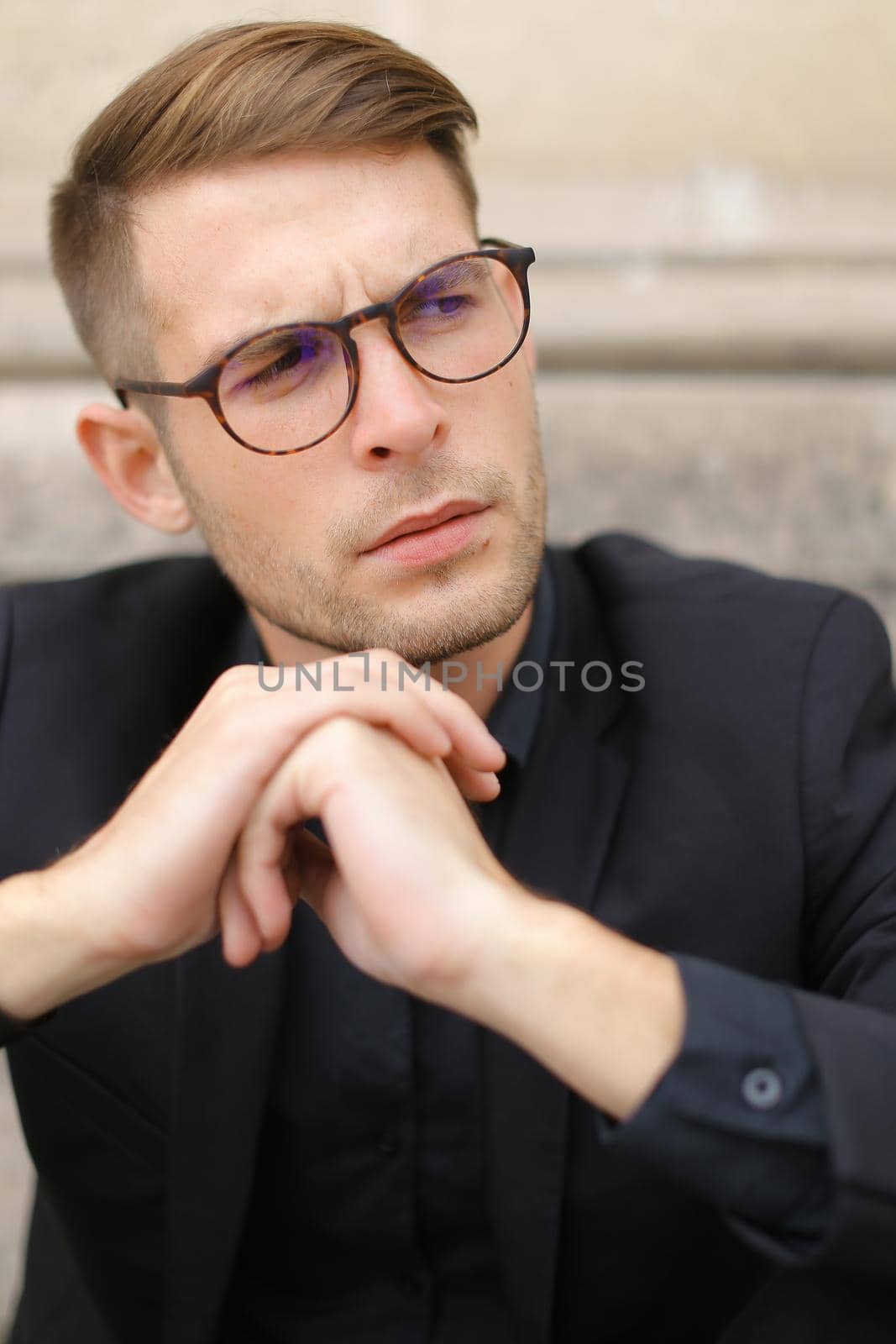 Closeup portrait of handsome man wearing black suit and glasses, sitting on sidewalk ground. Concept of male beauty and fashionable model.