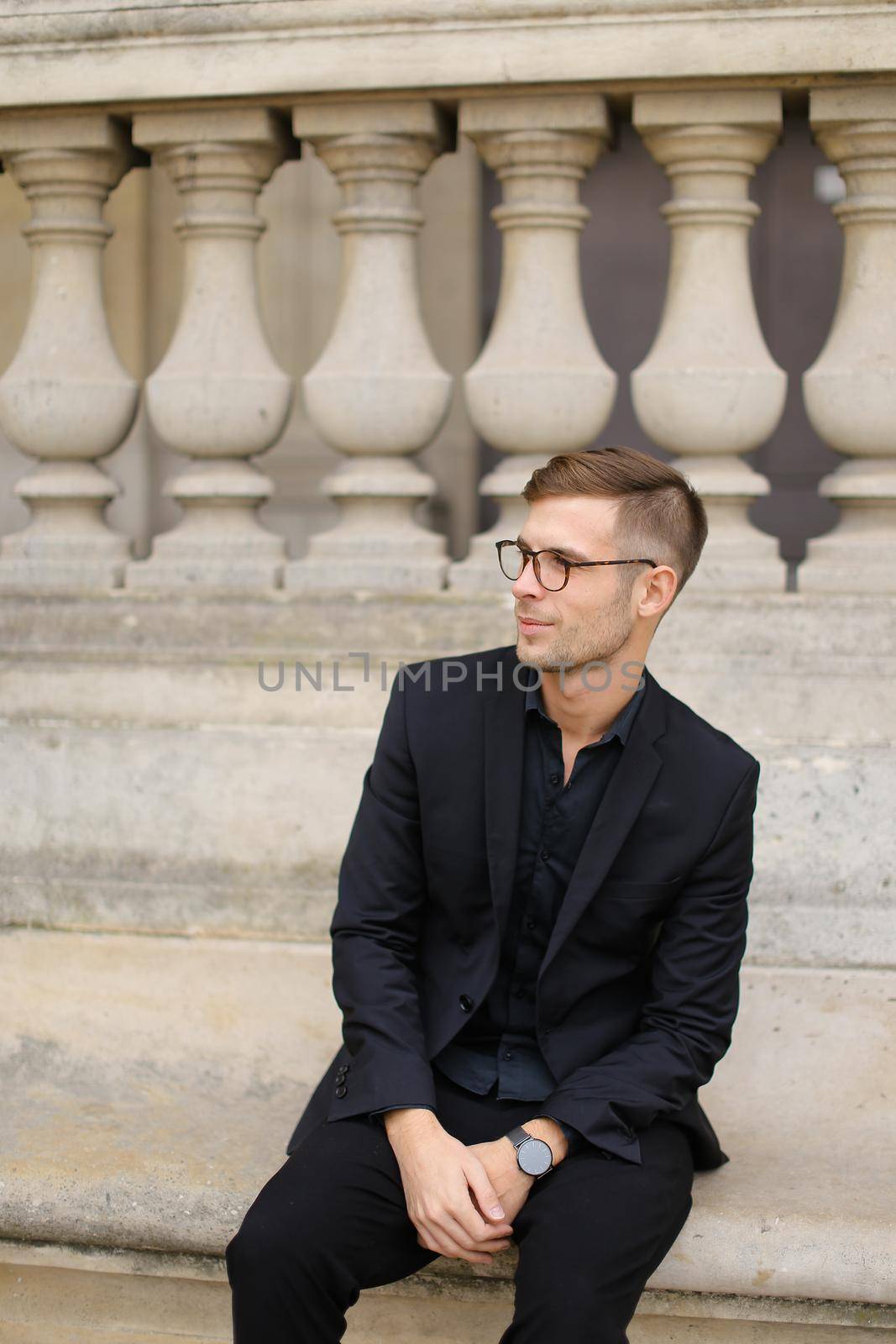 Young caucasian man sitting on sidewalk and leaning on concrete railing of building, wearing black suit and glasses. Concept of walkig in city, urban photo session and male person model.